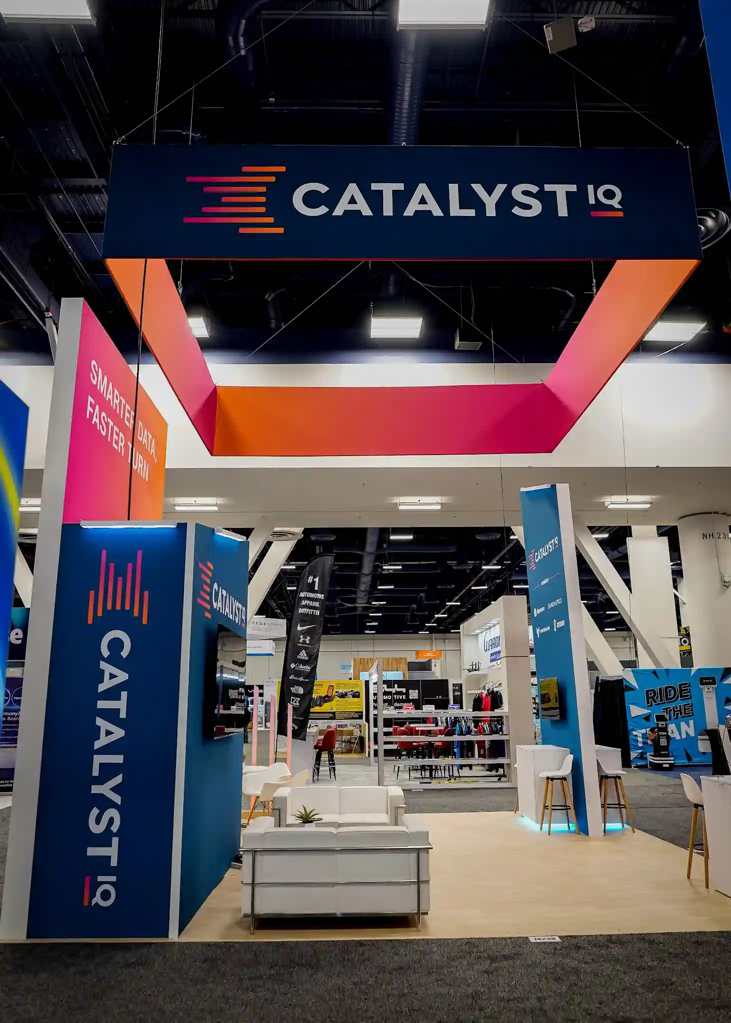 Catalyst IQ overhead hanging sign structure with teal and orange branding, viewed from the booth entrance showing lounge seating below