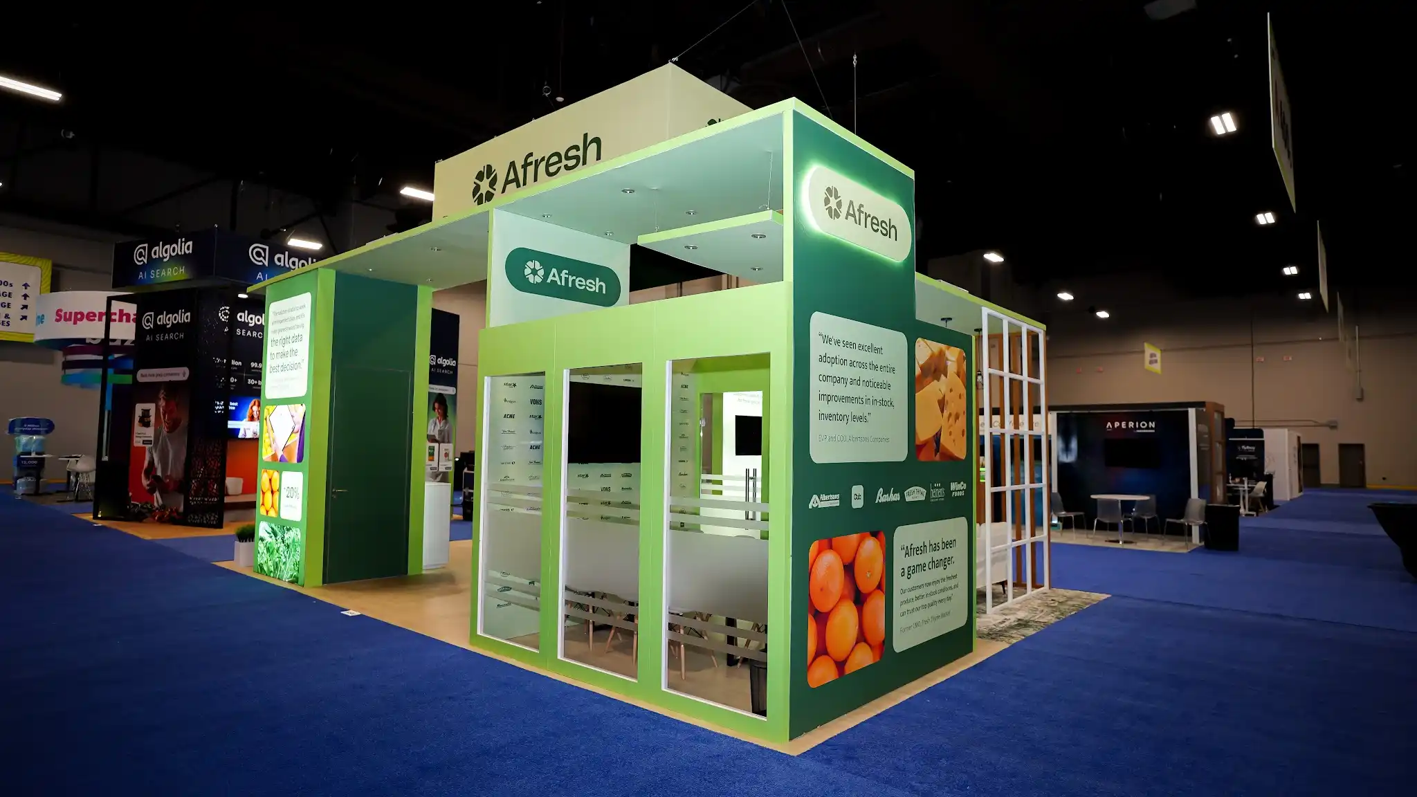 Back and side exterior view of the Afresh booth showing customer social proof wall with Albertsons WinCo Foods and Raleys logos, customer testimonial quotes, frosted glass meeting room visible behind, deep forest green panels, lime green accents