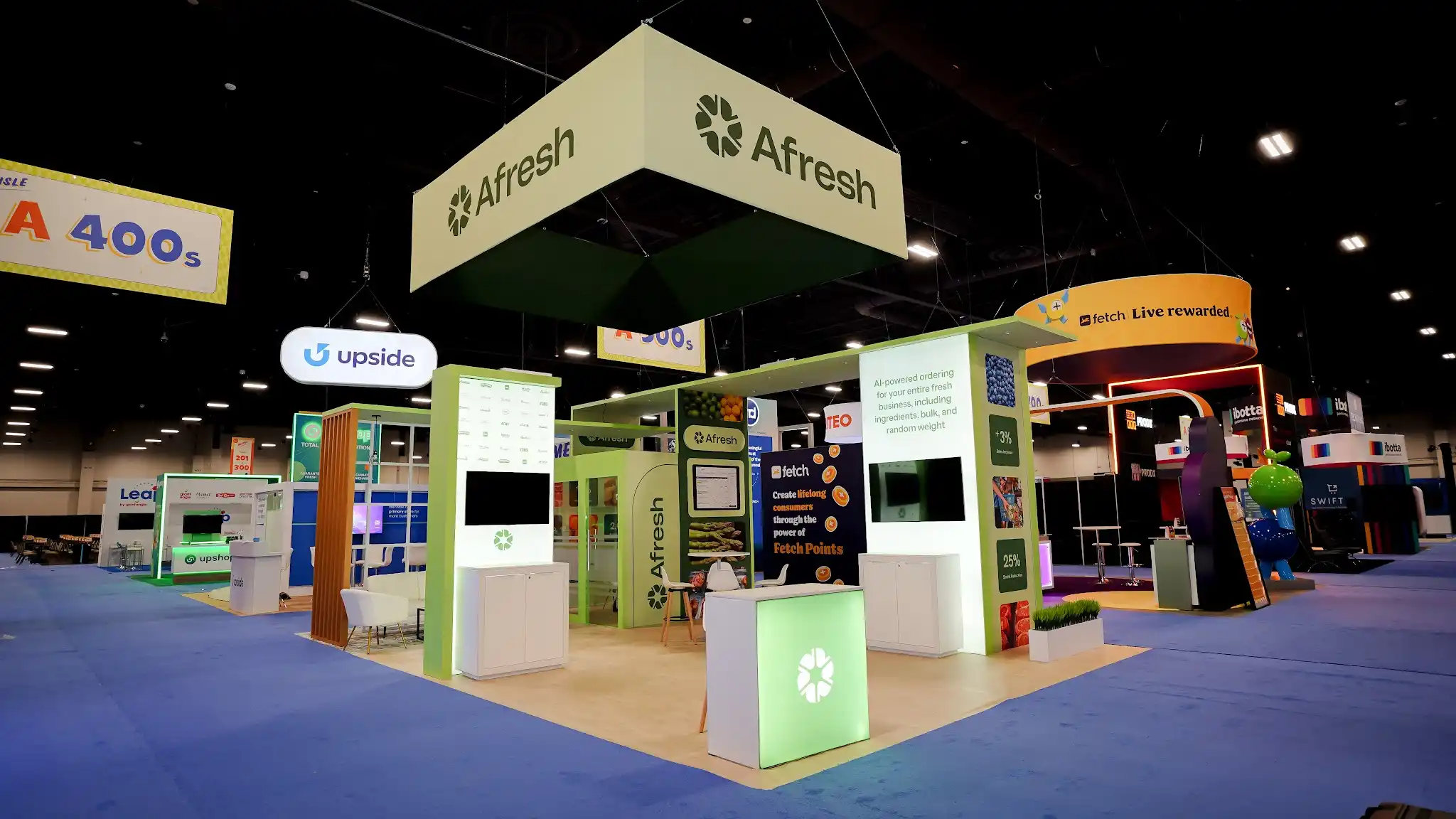 Wide-angle view of the Afresh booth at Groceryshop 2024 highlighting the illuminated LED logo cube emitting vivid lime-green light with white Afresh pinwheel logo at the booth entrance, interactive demo kiosks with flat-panel monitors visible in the interior, data storytelling columns with produce imagery and metrics, deep forest green panels, white bar stools, overhead ceiling-suspended Afresh sign, convention hall backdrop with neighboring exhibitor booths, overhead rigging and lighting