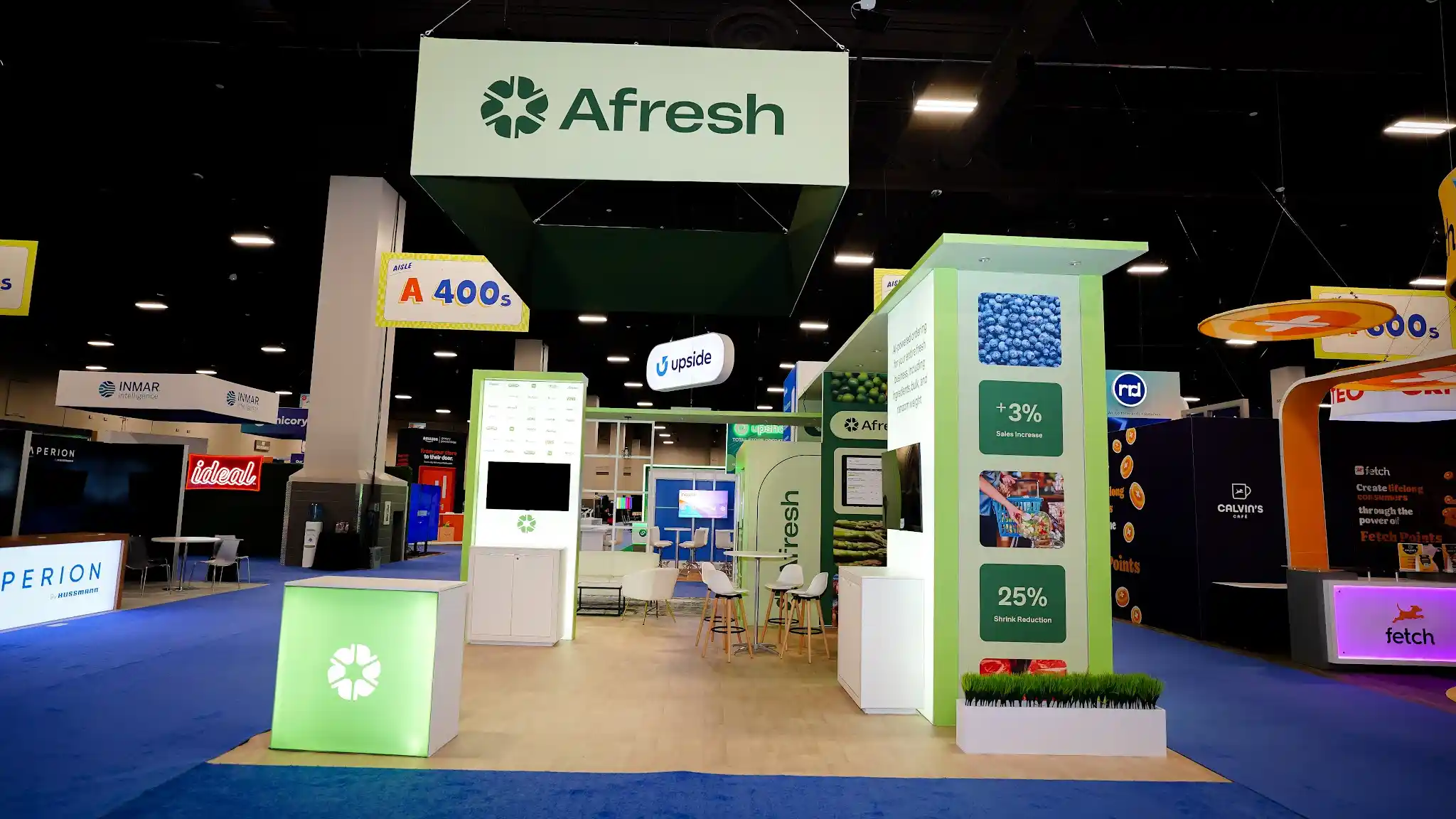 Wide establishing shot of the complete Afresh 20x30 ft island exhibit at Groceryshop 2024 in the Mandalay Bay Convention Center showing the front entry corner with two monumental data storytelling columns flanking the entrance, each layered with vivid produce photography of blueberries asparagus and citrus plus customer testimonial quotes and ROI metric tiles, large rectangular ceiling-suspended sign with cream face and dark green Afresh text and pinwheel logo, glowing lime-green LED beacon cube at the entrance, bright lime green structural frame and columns, deep forest green data panel backgrounds, white Scandinavian-style bar stools with wood legs, flat-panel demo monitors, natural wood flooring, green turf planters at column bases, neighboring exhibitor booths visible in background