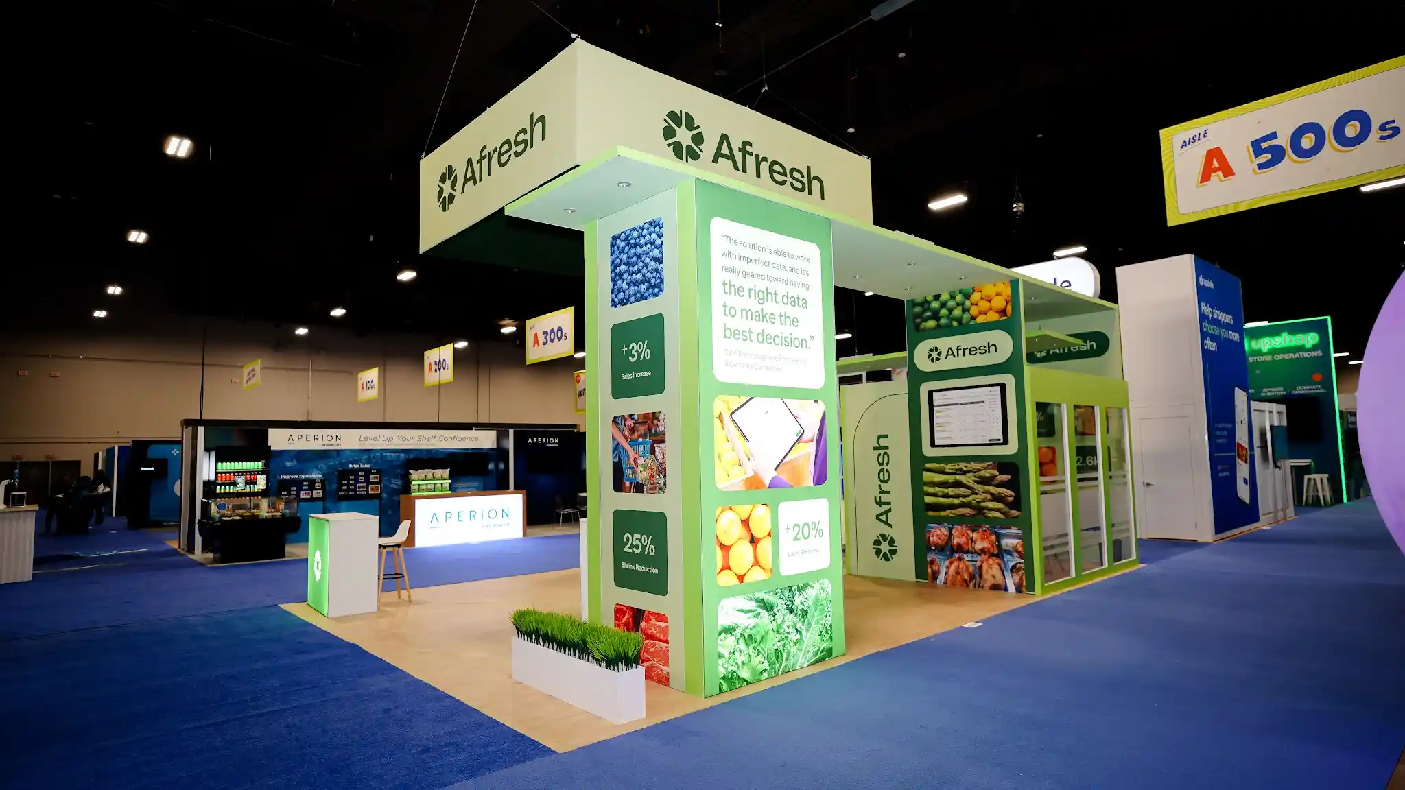 Front-left view of the Afresh 20x30 ft island booth at Groceryshop 2024 showing the data storytelling columns in detail with customer testimonial quote from Albertsons SVP and GM, ROI metric tiles showing plus 3 percent Sales Increase and 25 percent Shrink Reduction, vivid produce photography of blueberries and asparagus, customer logos including Albertsons WinCo Foods and Raleys, deep forest green data panel backgrounds, frosted glass meeting room visible in the background with green horizontal stripe accents, lime green structural frame elements, overhead ceiling-suspended Afresh sign