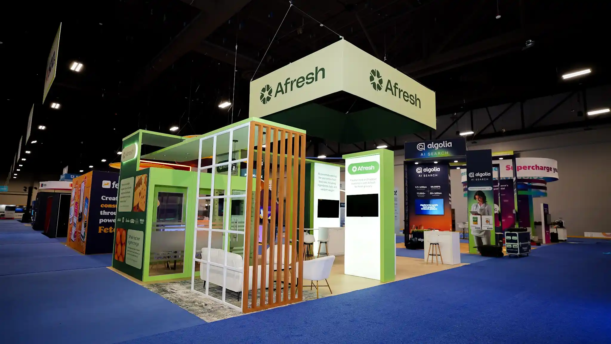 Front view of the Afresh 20x30 ft island booth at Groceryshop 2024 showing the warm honey-toned vertical wood slat partition creating a visual threshold to the hospitality lounge area, white upholstered lounge chairs and patterned area rug visible behind the slats, lime green structural columns, deep forest green data panels with produce photography and metrics, overhead Afresh ceiling sign, white bar stools at counter-height tables, flat-panel monitors, green turf planters, natural wood flooring, Mandalay Bay Convention Center environment