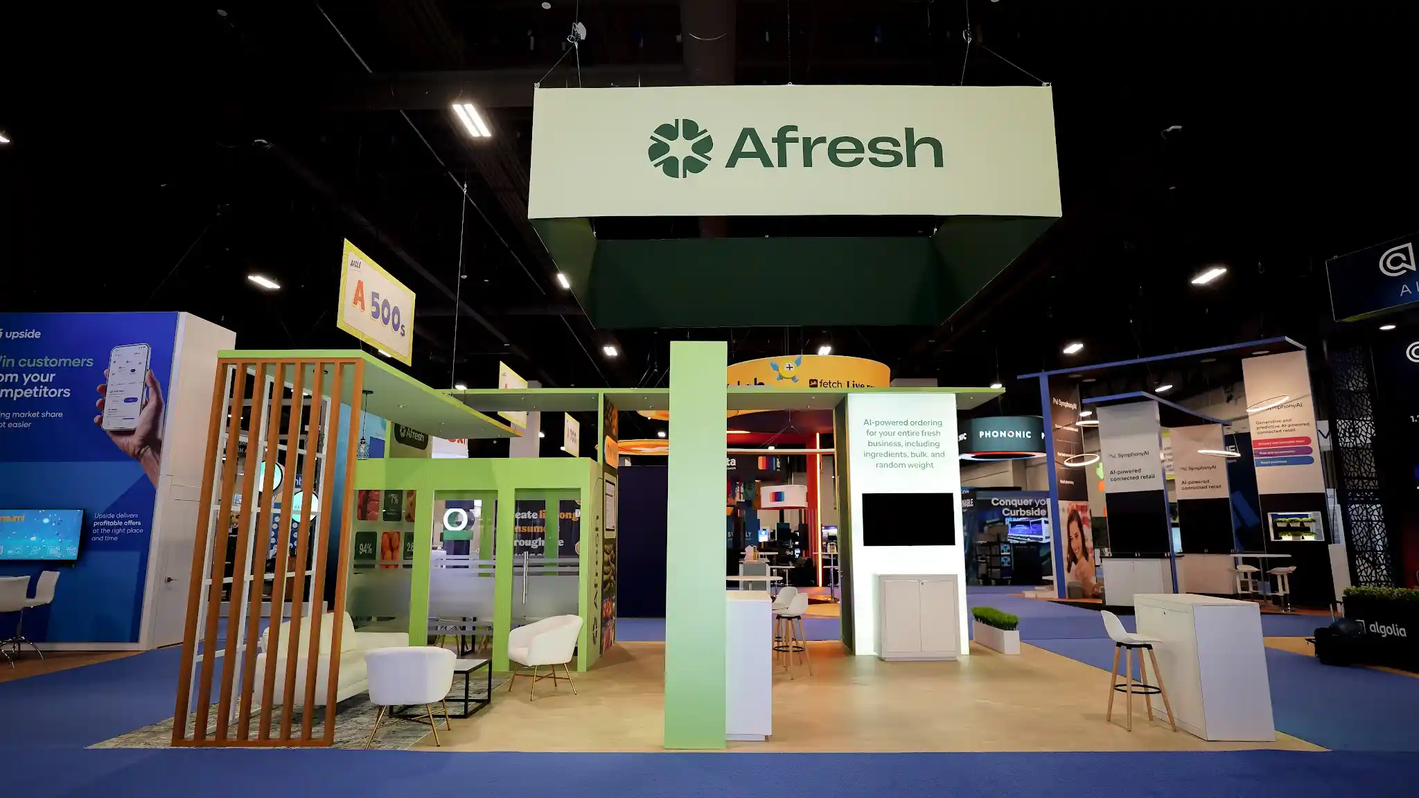 Full-width frontal view of the Afresh booth at Groceryshop 2024 showing the complete booth architecture with three structural columns in bright lime green, product positioning panel reading AI-powered ordering for your entire fresh business including ingredients bulk and random weight, deep forest green data panel backgrounds with customer logos and ROI metrics, white surfaces throughout, overhead Afresh ceiling sign, flat-panel demo monitors, green turf planters, natural wood flooring