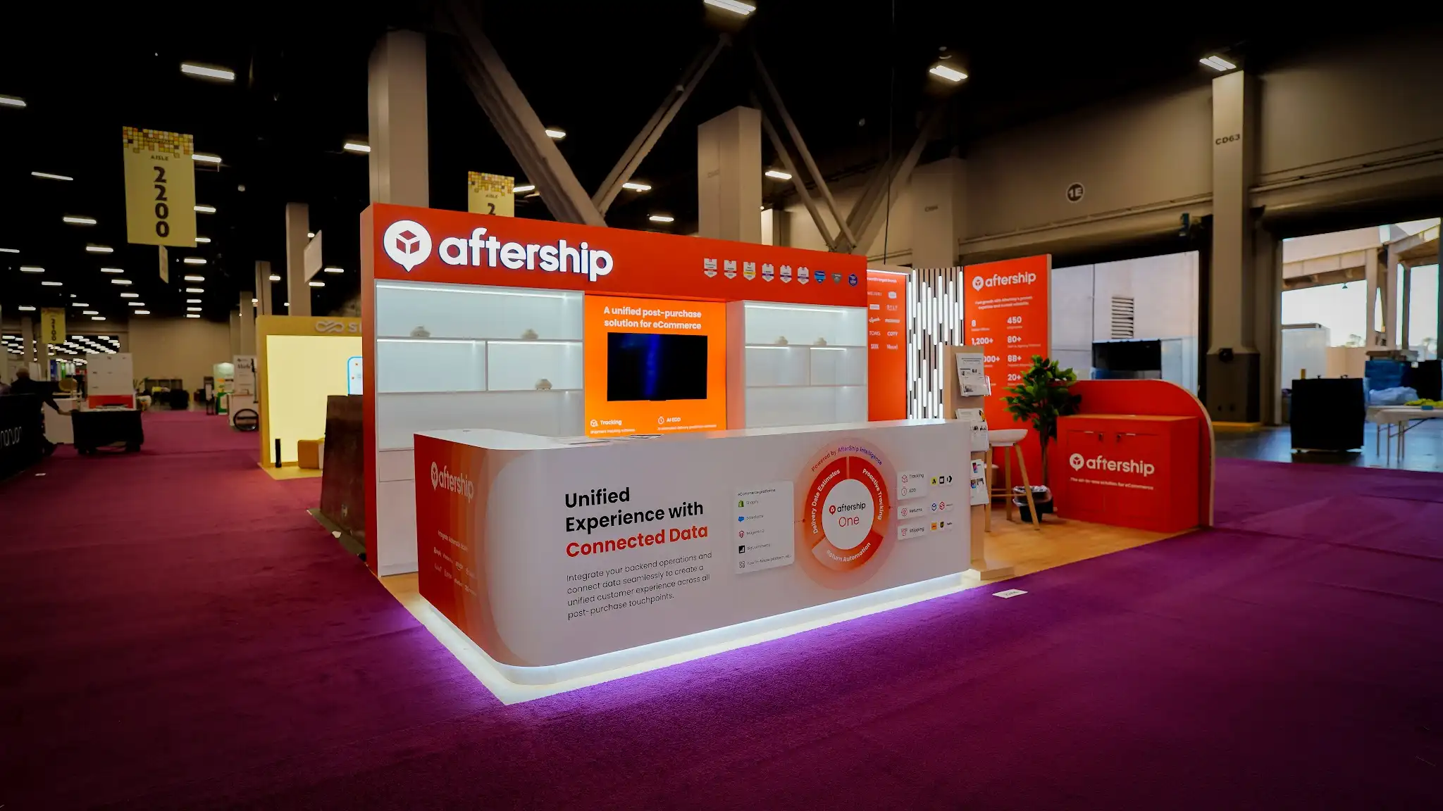Front-left angle view of the AfterShip booth at Shoptalk 2025 showing the curved white reception counter with purple-pink LED underglow displaying AfterShip One circular platform diagram and Integrate Automate Scale tagline with Shopify TikTok Shop Amazon partner logos, bold orange header fascia with AfterShip logo and award badges, illuminated white modular shelving, central orange media panel with flat-screen monitor, company statistics panel showing 8 Global Offices 450 Employees 1200+ Carriers 20000+ Paid Customers 8B+ Tracked Shipments, green tropical plant in corner, light wood laminate flooring, deep magenta convention carpet