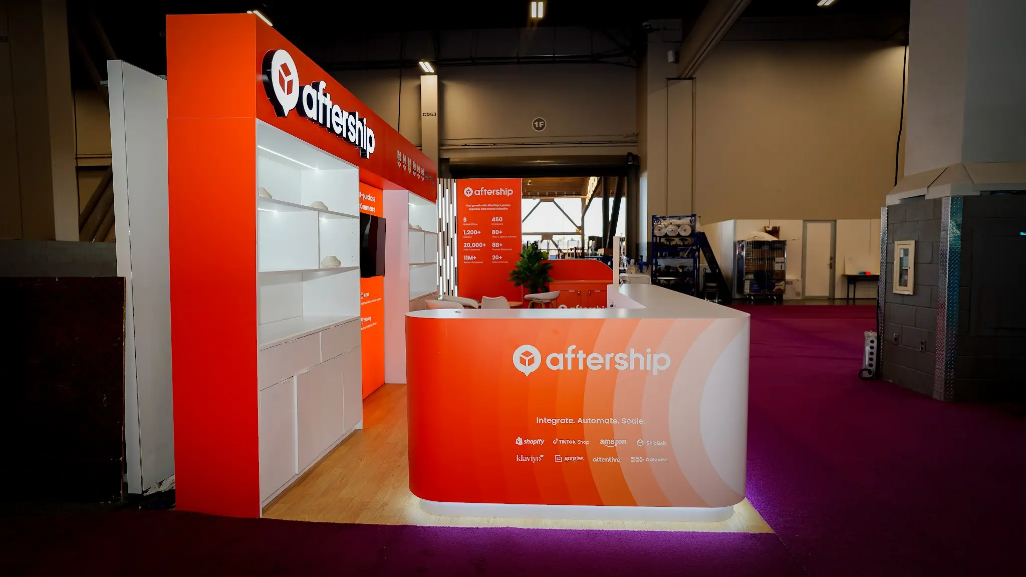 Right side exterior view of the AfterShip booth showing orange statistics tower, white illuminated shelving, flat-screen monitor, curved reception counter with Integrate Automate Scale tagline and partner logos, green tropical plant, light wood flooring, deep magenta convention carpet, TOPSOFT booth visible in background