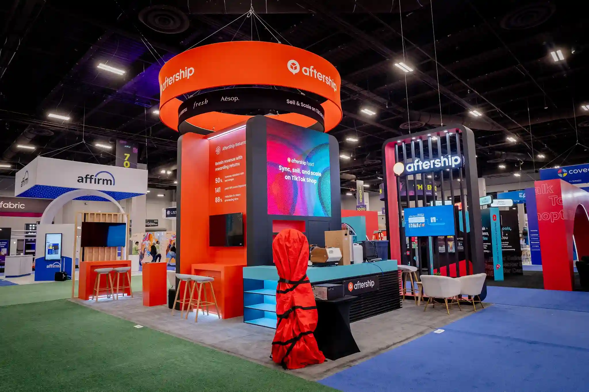 AfterShip 20x30 custom island booth at Shoptalk 2024 featuring large LED video wall screens displaying product demos, bold orange and black brand colors, sculptural hanging sign structure, open meeting areas, and dramatic overhead lighting at Booth 630 in the Mandalay Bay Convention Center in Las Vegas