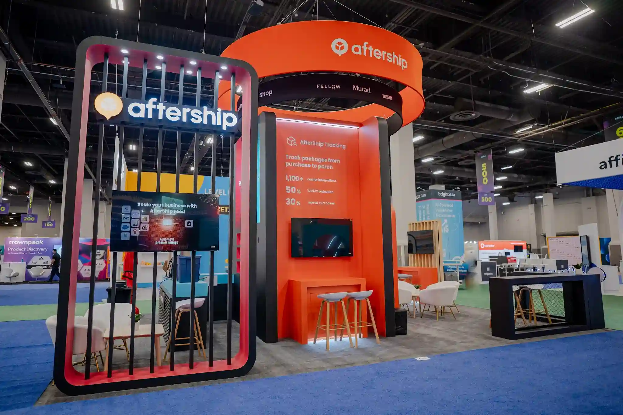 Wide angle view of AfterShip 20x30 island booth at Shoptalk 2024 showing the complete architectural structure with orange and black color scheme, open pavilion layout, LED video walls, sculptural hanging sign, meeting areas with branded furniture, and dramatic overhead lighting creating a bold presence on the Mandalay Bay expo floor
