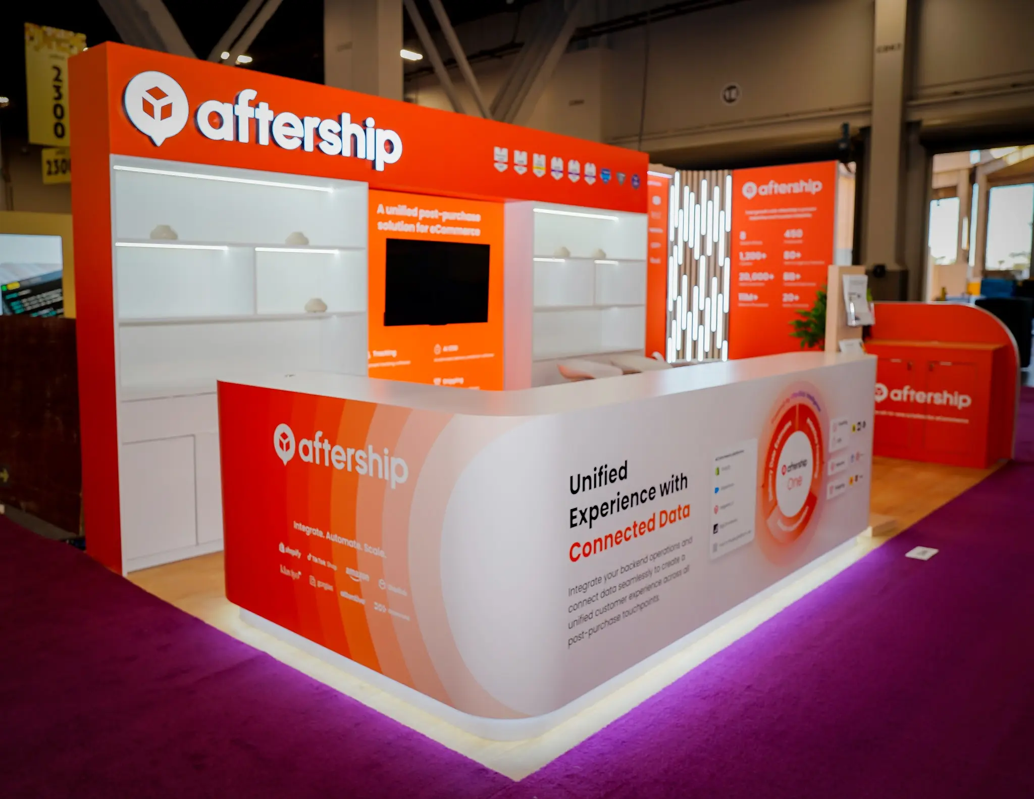 Low-angle close-up perspective of the AfterShip booth at Shoptalk 2025 showing the curved white reception counter with AfterShip logo on red-orange gradient, Unified Experience with Connected Data text in bold black, AfterShip One circular platform diagram in red and white, integration partner logos Shopify TikTok Shop Amazon Klaviyo Gorgias Attentive ShipBob, purple-pink LED underglow on deep magenta carpet, orange back wall with illuminated white shelving, central media panel with A unified post-purchase solution tagline and large monitor, Tracking AI EDD Shipping service icons, orange header fascia with AfterShip wordmark and award badges