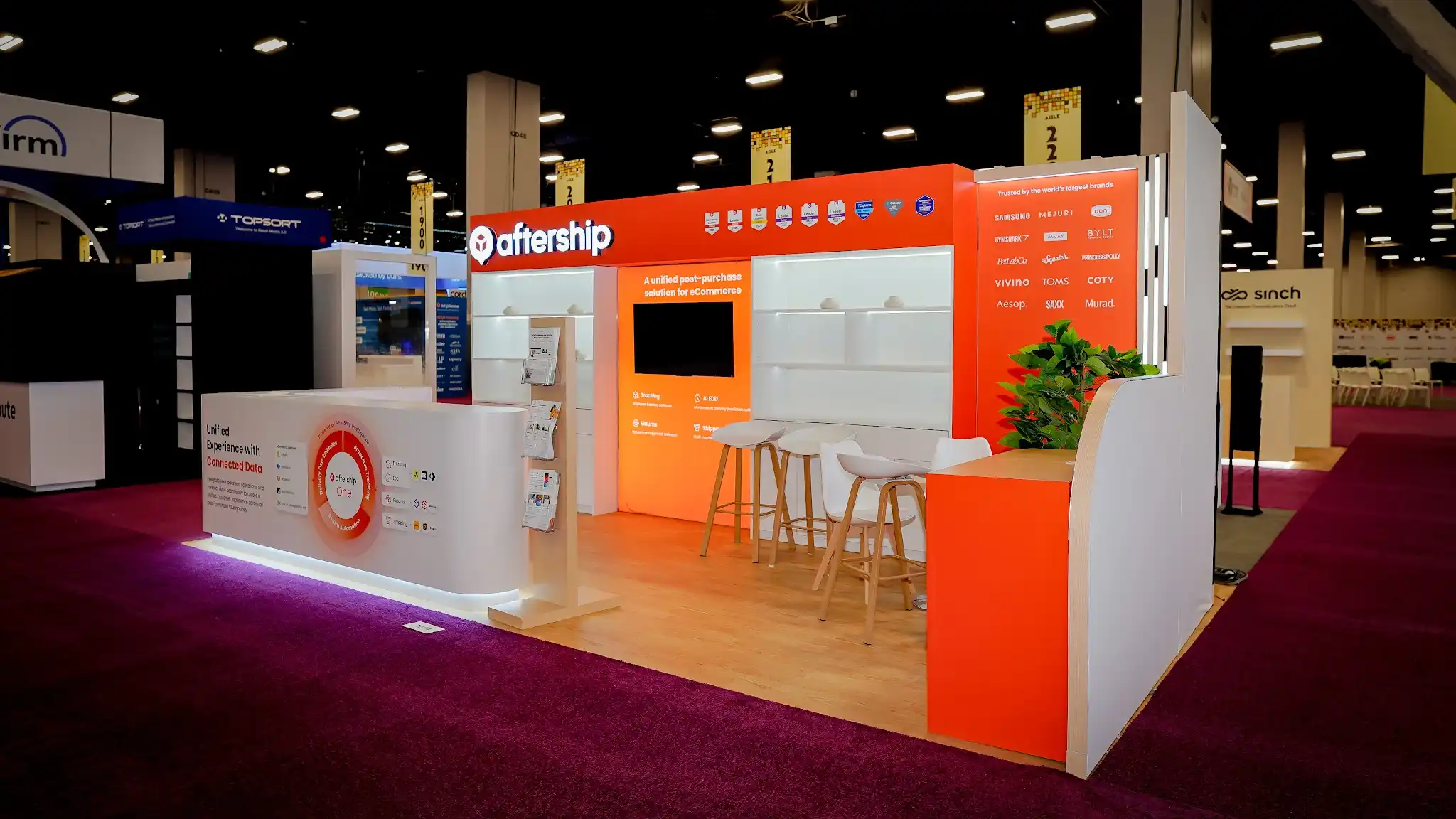 Wide front-right perspective of the AfterShip 10x20 ft booth at Shoptalk 2025 capturing the full width of the exhibit including the orange branded header with AfterShip logo and award badges, white illuminated modular shelving units, central orange media panel with A unified post-purchase solution tagline and flat-screen monitor, Trusted by the worlds largest brands panel with Samsung Mejuri Ooni Gymshark BYLT PetLabCo Squatch Princess Polly Vivino TOMS COTY Aesop SAXX Murad logos in white on orange, vertical LED light column, curved white reception counter with partner logos, orange curved lounge-meeting area in background, green tropical plants, light wood laminate flooring, white bar stools with wood legs