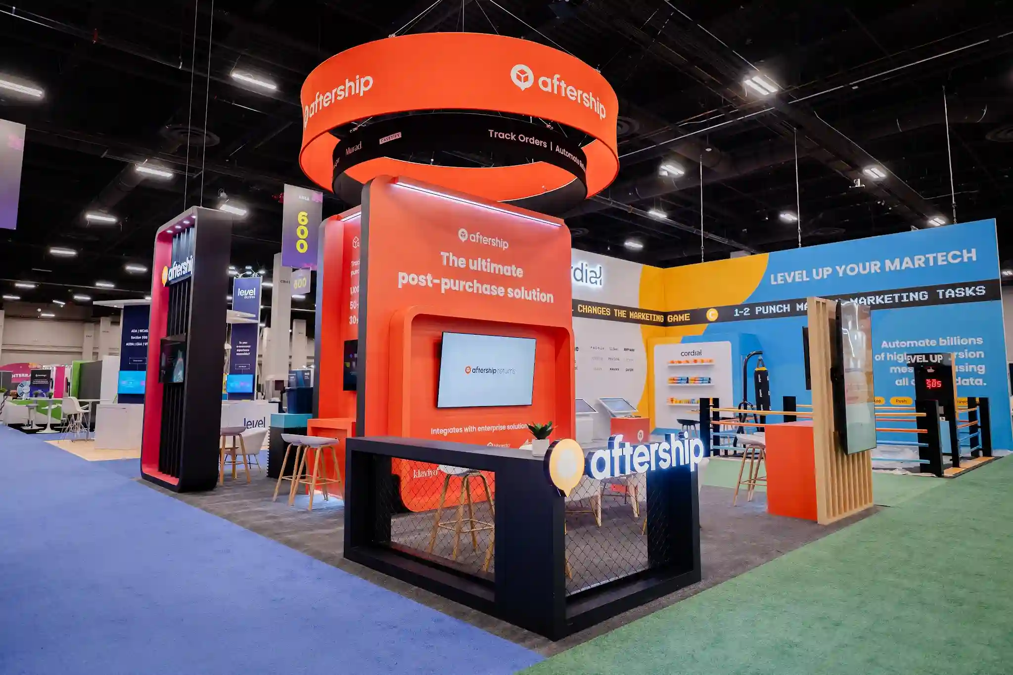 AfterShip island booth showing bold orange floor-to-ceiling panels, black structural framework, open meeting areas with branded orange furniture, LED screens displaying product demos, and overhead signage structure at Shoptalk 2024 Mandalay Bay Convention Center