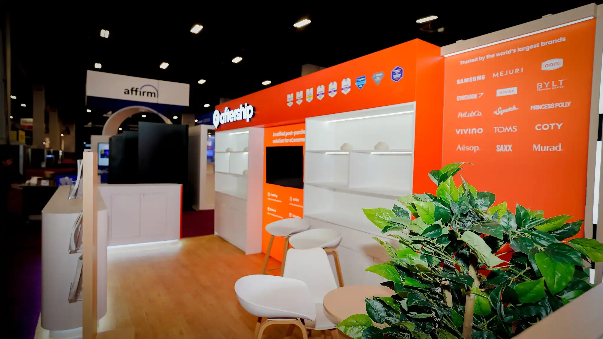Interior close-up of the AfterShip booth at Shoptalk 2025 showing brand trust wall with Samsung Mejuri Ooni Gymshark BYLT logos in white on orange, illuminated white shelving, flat-screen monitor, product service icons, white modern chairs with wood legs, green tropical plant in foreground, Affirm booth visible in background