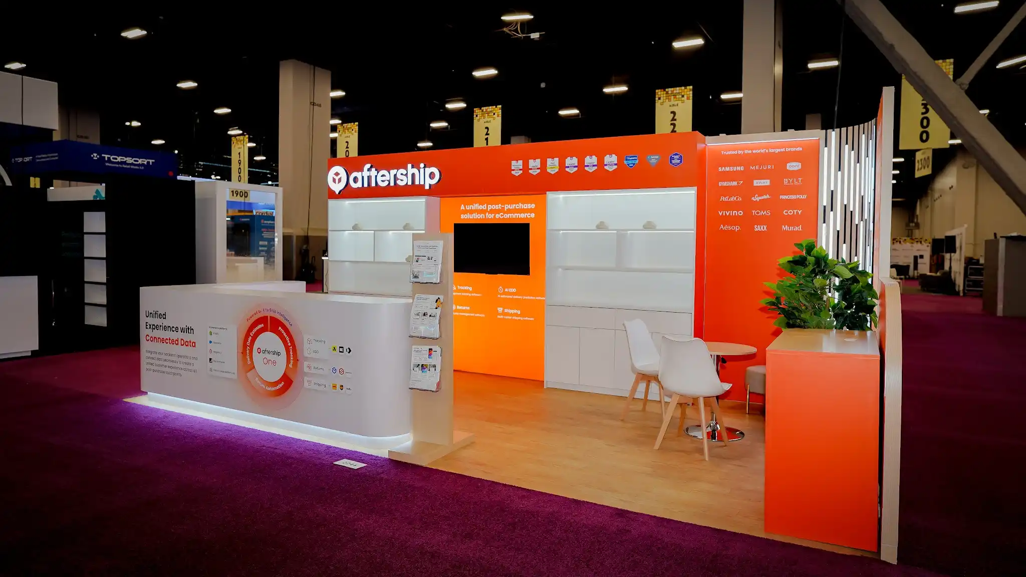 Wide front-facing establishing shot of the complete AfterShip 10x20 ft custom island booth at Shoptalk 2025 Booth 2244 at Mandalay Bay Convention Center Las Vegas showing the full width of the orange and white branded back wall with AfterShip logo and wordmark in white on a bold orange header fascia, row of industry award badge icons, illuminated white modular shelving, central orange media panel reading A unified post-purchase solution for eCommerce with large flat-screen monitor, Trusted by the worlds largest brands panel with Samsung Gymshark TOMS Aesop and other brand logos in white on orange, vertical LED light column with individual white LED bars, curved white reception counter with purple-pink LED underglow displaying AfterShip One circular platform diagram and Integrate Automate Scale tagline with Shopify TikTok Shop Amazon Klaviyo Gorgias Attentive ShipBob partner logos, light wood laminate flooring, green tropical plants, white modern Eames-style chairs, deep magenta convention hall carpet, neighboring exhibitor booths in background