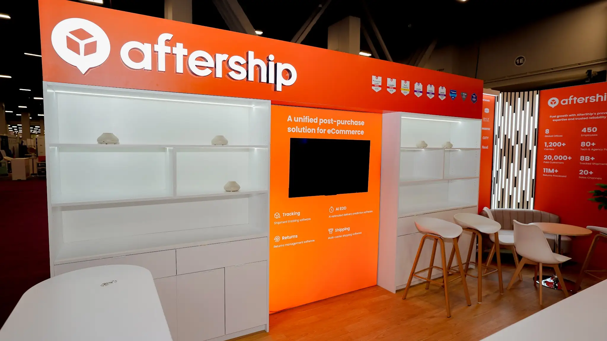 Straight-on close-up photograph of the AfterShip booth back wall at Shoptalk 2025 showing detailed view of the orange header fascia with AfterShip logo white cube icon in speech bubble and aftership wordmark in large white bold text, row of approximately 8 to 10 industry award certification badge icons in blue silver gold, illuminated white modular shelving with soft internal lighting and small white rounded decorative objects, central orange content panel reading A unified post-purchase solution for eCommerce above large flat-panel monitor, four service offering icons in 2x2 grid for Tracking AI EDD Returns and Shipping in white on orange, Trusted by the worlds largest brands headline with Samsung Mejuri Ooni Gymshark BYLT PetLabCo Squatch Princess Polly Vivino TOMS COTY Aesop SAXX Murad logos in white on orange, tall vertical LED light column with white LED bars, company statistics panel with 8 Global Offices 450 Employees 1200+ Carriers numbers in white on orange