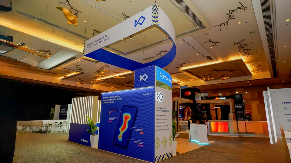 Dramatic wide-angle low-vantage exterior shot of the Sardine booth at MRC Vegas 2026 emphasizing the curved white-and-blue ribbon hanging sign towering above the booth with underside text clearly reading Agentic Platform to Fight Fraud and Abuse with Sardine angular diamond logo icon and yellow-and-black chevron accents at ends suspended via black rigging cables, navy-blue wall panels with pixel-dot texture below displaying Agentic platform text and Sardine logo, graphic panel showing smartphone with heat-map visualization for bot-driven carding attack detection, five-bullet unified platform list, curved white reception counter with blue LED base glow branded Sardine in white, warm golden amber hotel ballroom lighting creating dramatic contrast with brightly lit booth, neighboring Mastercard Southwest Airlines Nu Skin Autotrader Wyndham Hotels booths visible in background, decorative gold fish bird ceiling sculptures, dark gray convention carpet