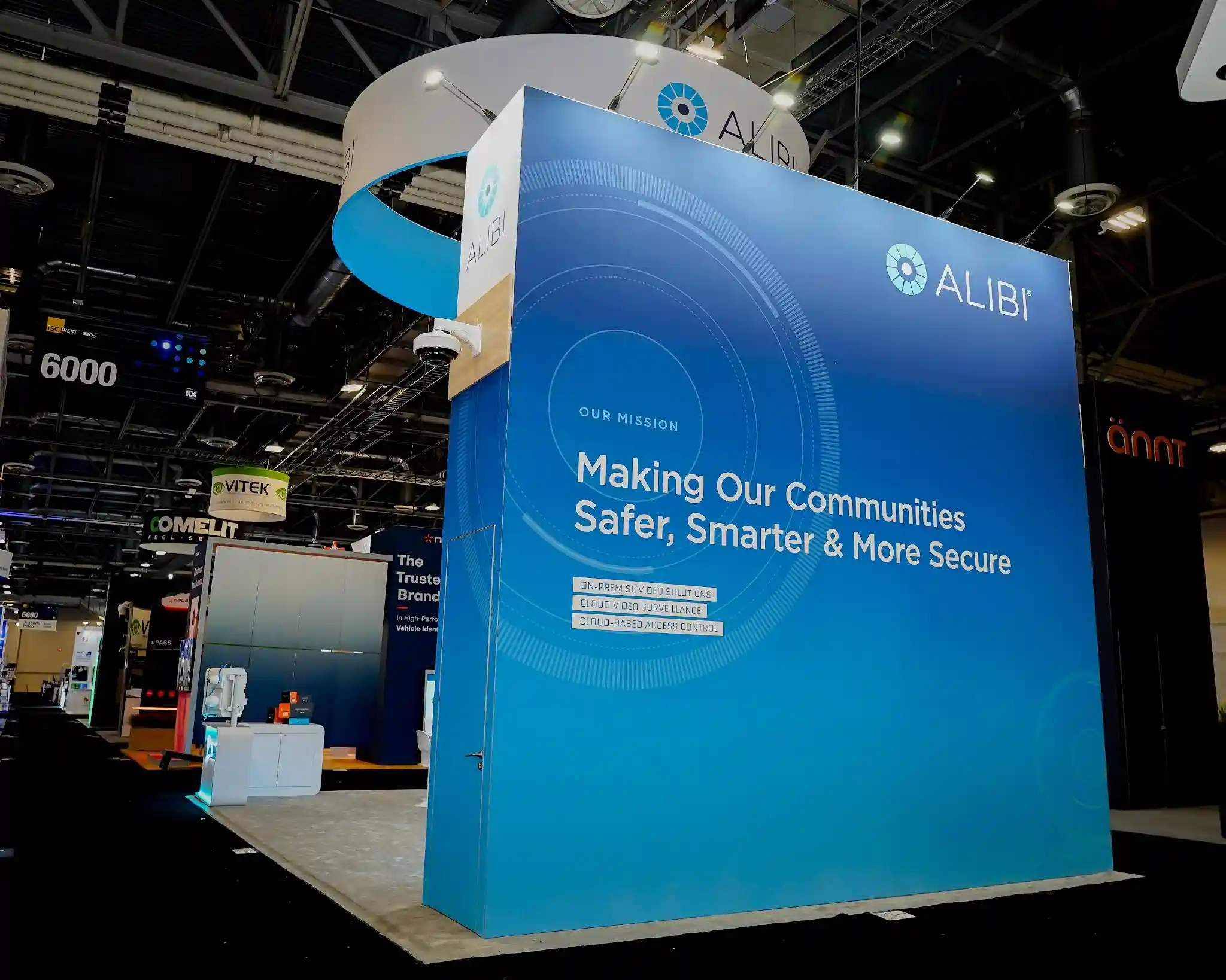 Alibi Security exterior mission wall featuring deep blue background with concentric circular radar pattern, large Alibi logo with gear icon, Our Mission Making Our Communities Safer Smarter and More Secure headline, On-Premise Video Solutions Cloud Video Surveillance and Cloud-Based Access Control listed below, security camera mounted at top corner, and circular hanging sign visible above