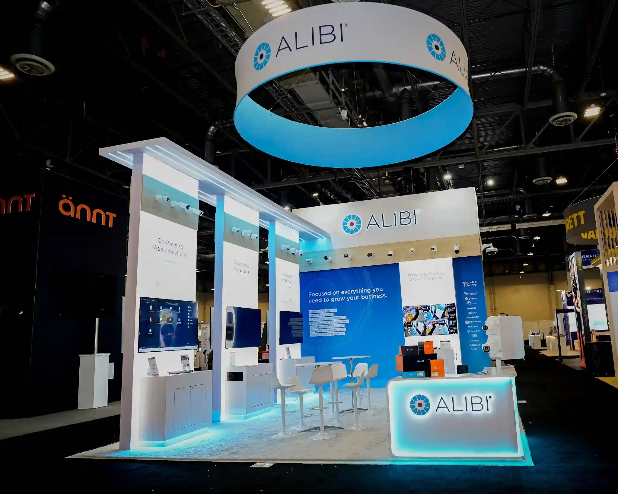 Reception perspective showing backlit Alibi logo on front counter, circular overhead sign, blue LED ambient glow, 16-camera grid monitor, and white bar stools with integrated booth lighting