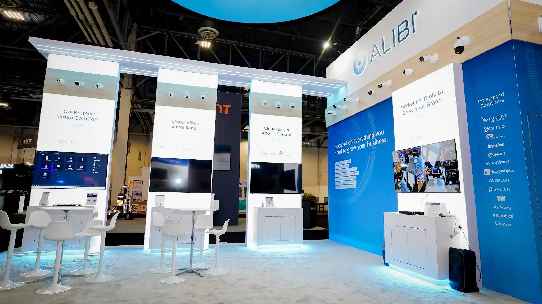 Three demonstration bays with tall white pillars, headers reading On-Premise Video Solutions Cloud Video Surveillance and Cloud-Based Access Control, widescreen monitors showing security feeds and dashboards, blue Integrated Solutions partner pillar listing Eagle Eye Brivo and Milestone, security cameras mounted above screens, and blue ambient floor glow