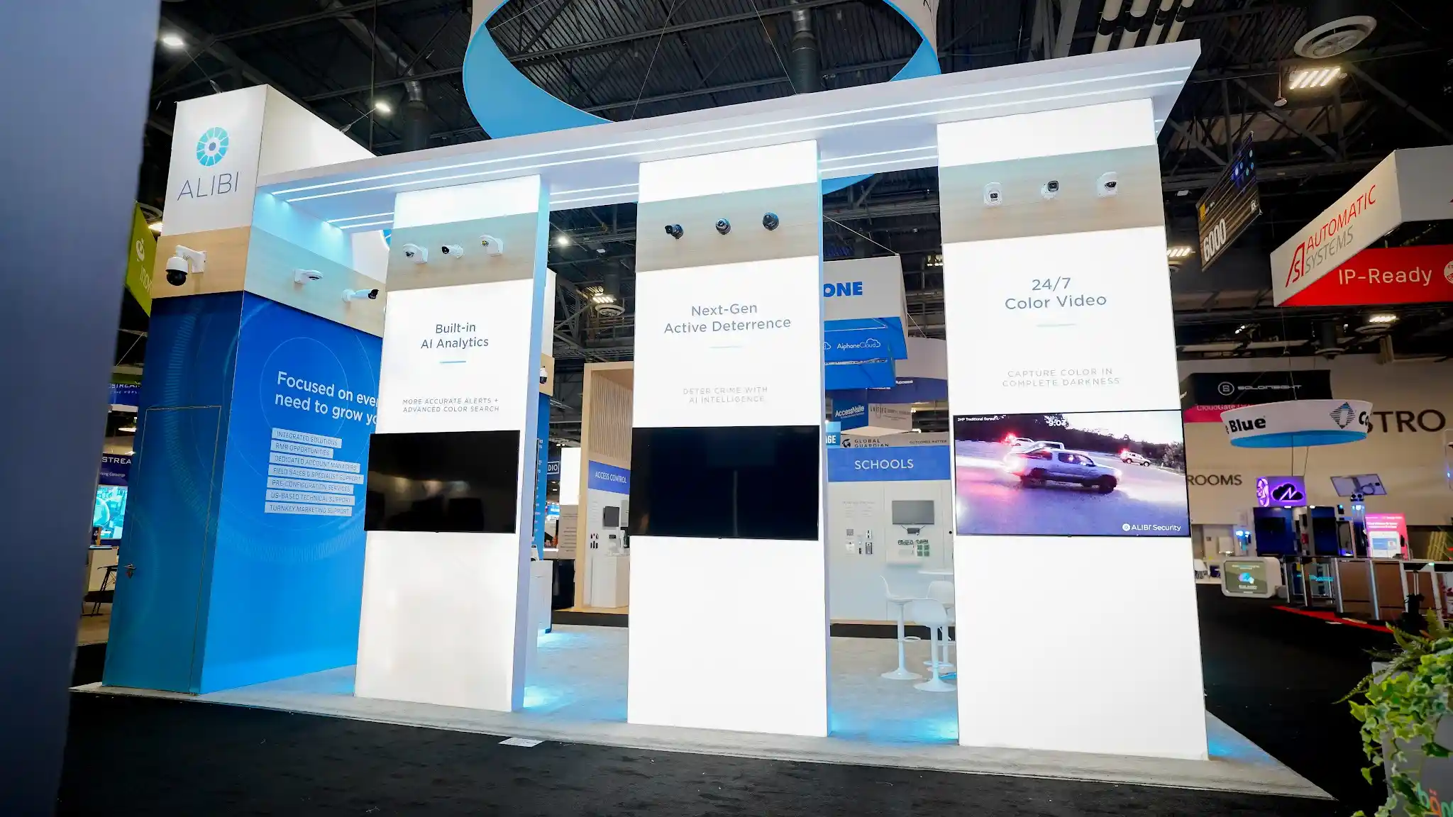 Four informational panels with headers Built-in AI Analytics Next-Gen Active Deterrence and 24/7 Color Video, corresponding security cameras mounted above each panel, widescreen monitors showing nighttime vehicle footage and AI interface, Focused on everything you need to grow your business central messaging, and white structural elements with blue accent wall