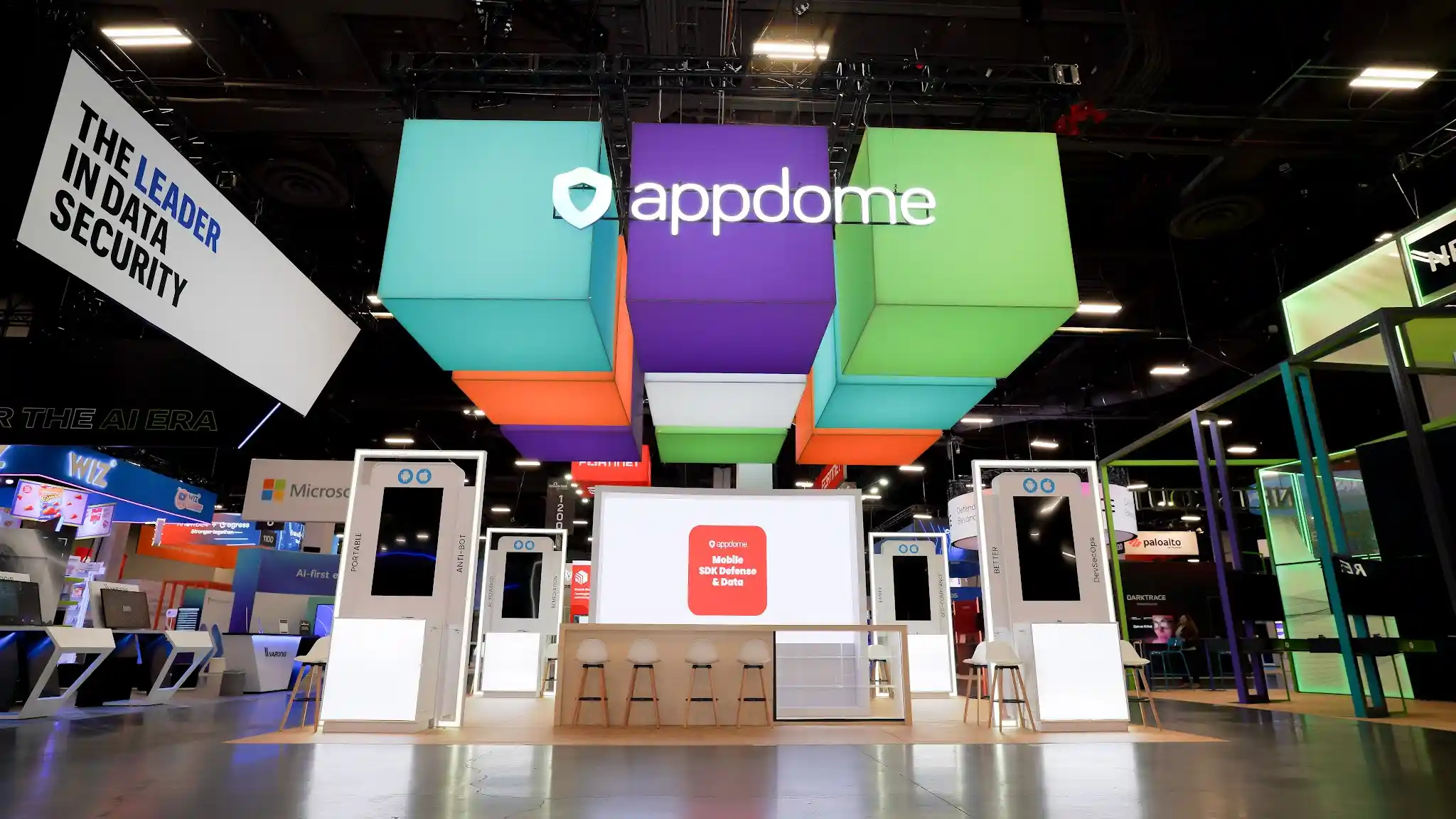 Direct frontal view of Appdome island exhibit showing brand name spelled across colorful hanging cubes in purple turquoise lime green coral orange and white, symmetric white MOBILE LINK kiosk placement on both sides, central meeting area with natural wood bar-height stools, orange display panel with product messaging, natural wood flooring throughout, and warm inviting material palette contrasting with the technical cybersecurity environment at Booth 1200 Black Hat USA 2024