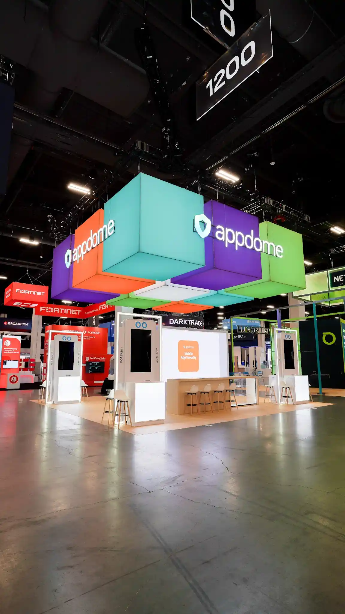 Angular aisle view of Appdome island exhibit at Black Hat USA 2024 showing Booth 1200 signage, colorful cube cluster sculpture overhead in purple turquoise lime green and coral orange, white kiosk stations with clean modern profiles, natural wood flooring and bar stools, adjacent Fortinet booth visible providing scale context for the vibrant Appdome island on the Mandalay Bay Convention Center Business Hall floor