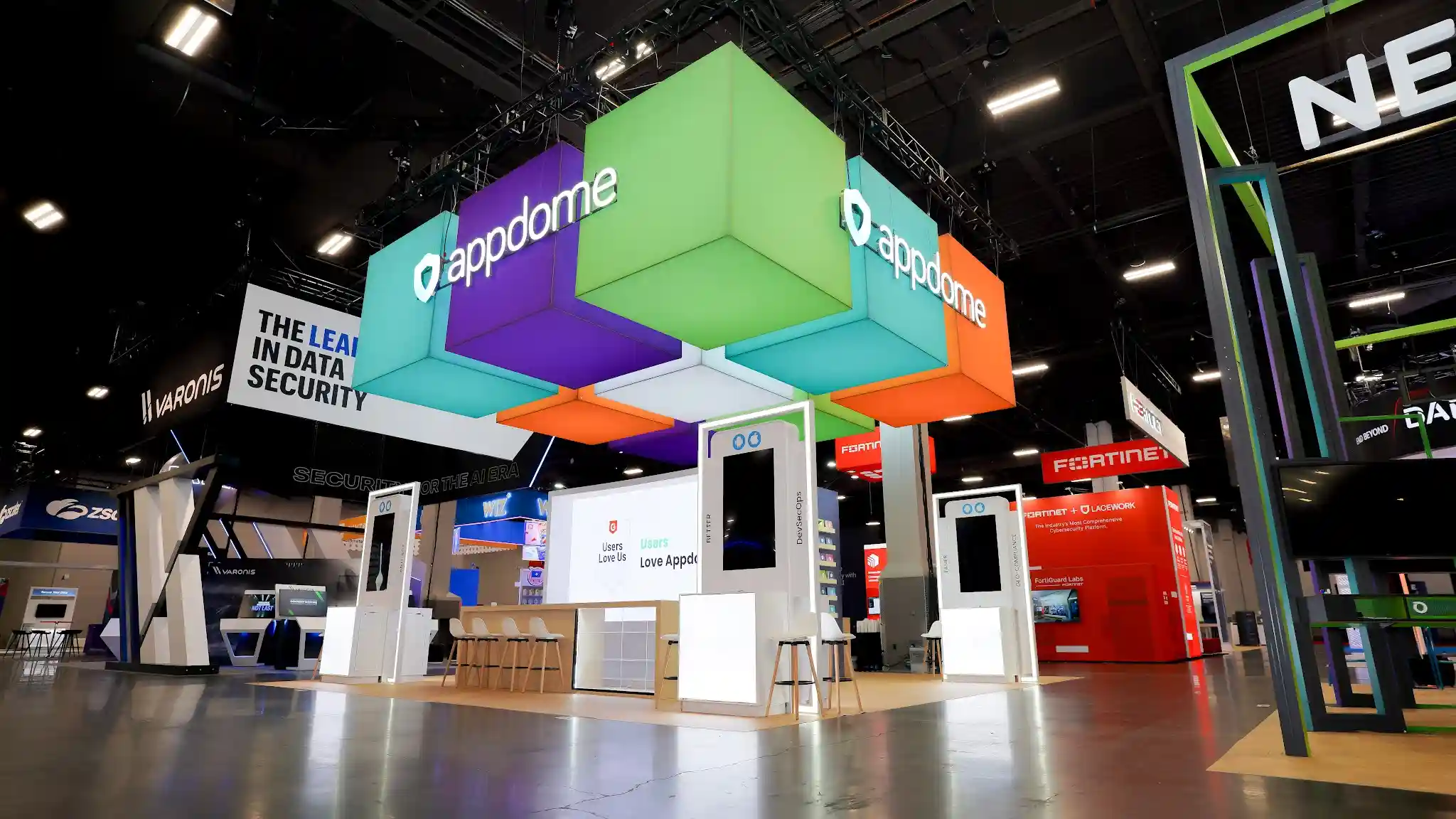 Wide angle side diagonal view of Appdome 30x30 island exhibit at Black Hat USA 2024 showing the booth commanding presence on the show floor with colorful suspended cube sculpture visible from distance, Users Love Us messaging on display panels, white kiosk stations and wood flooring creating warm inviting atmosphere, neighboring Varonis and Fortinet cybersecurity booths visible for scale context, and open four-aisle island access at Booth 1200 Mandalay Bay Convention Center