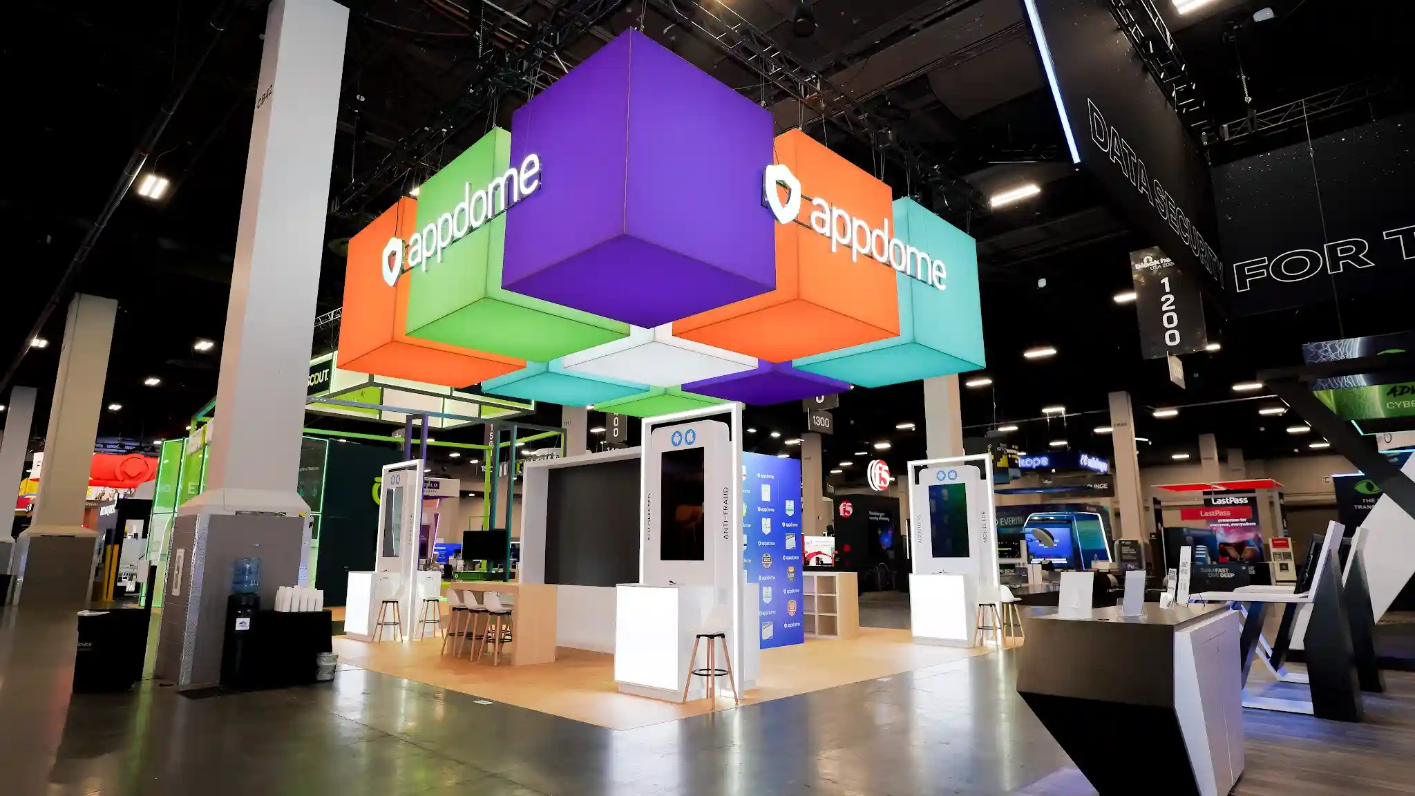 Wide-angle view of Appdome island exhibit at Black Hat USA 2024 showing the booth in context with neighboring cybersecurity booths on the Business Hall floor, colorful hanging cube sculpture in purple turquoise lime green and coral orange visible from distance as a wayfinding landmark, white kiosk stations and natural wood flooring contrasting with the dark corporate booths surrounding the island exhibit