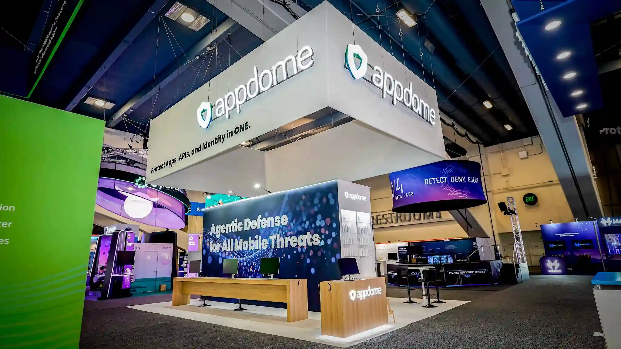 Wide dramatic right-front establishing shot of the complete Appdome 20x30 ft island exhibit at RSA Conference 2026 Booth 754 in the South Hall of Moscone Center San Francisco showing the massive white internally-illuminated rectangular hanging sign suspended by steel cables from the convention ceiling with the Appdome shield logo in teal green and appdome wordmark on the front face and Protect Apps APIs and Identity in ONE tagline visible on the right side face, the commanding dark navy backwall approximately 20 feet wide by 10 feet tall displaying Agentic Defense for All Mobile Threats in large bold white text over a particle bokeh constellation graphic of hundreds of soft glowing blue teal and white orbs, a sleek white branded podium in the right foreground with the internally illuminated Appdome shield logo glowing on its face, a natural wood bench approximately 6 to 8 feet long in light Scandinavian style sitting at center of the white raised platform floor, a thin cyan teal LED accent strip running along the top edge of the dark navy backwall creating a halo cove light effect, white aluminum structural framing, demo screens visible in the back area, neighboring Y4 Data Labs booth with curved LED screen showing DETECT DENY EJECT visible to the right, JFrog bright neon green booth partially visible to the left, dark charcoal convention hall carpet surrounding the white platform, industrial Moscone Center South Hall ceiling with rigging and track lighting overhead