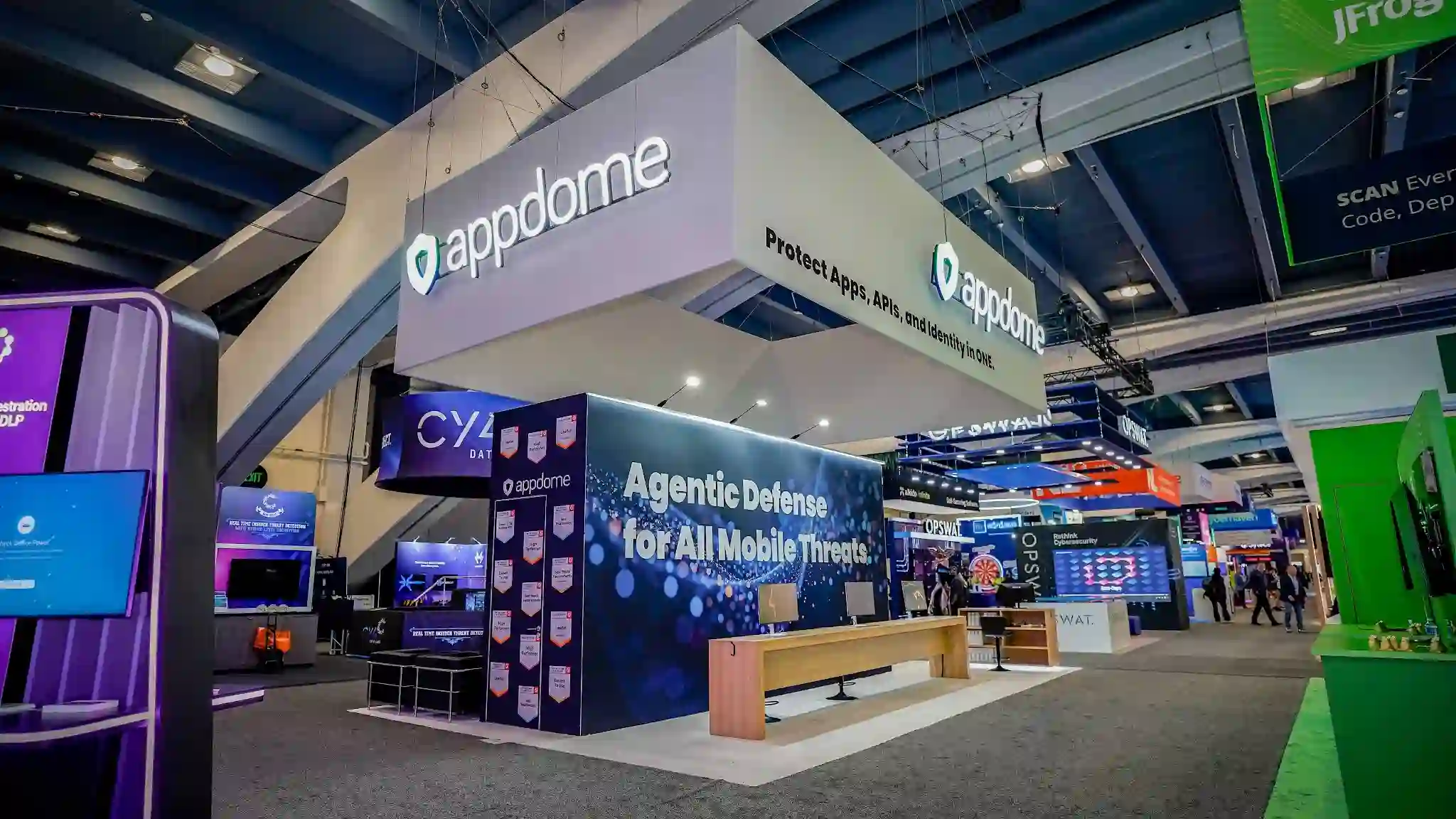 Wide front-left establishing shot of the complete Appdome 20x30 ft island exhibit at RSA Conference 2026 taken from approximately 20 to 25 feet away at a low angle showing the full height and breadth of the booth with the massive white internally-illuminated rectangular hanging sign centrally positioned overhead suspended by steel cables with appdome wordmark and teal green shield logo visible on the front face and Protect Apps APIs and Identity in ONE tagline on the right side face, the commanding dark navy backwall module approximately 20 feet wide by 10 feet tall displaying Agentic Defense for All Mobile Threats in large bold white text over the particle bokeh constellation graphic of glowing blue teal and white orbs, the natural light wood Scandinavian bench sitting at center of the white raised platform floor, demo stations with two medium flat screens in the back-left area alongside black metal bar stools, the tall freestanding right-side tower column with Appdome logo and G2 awards badges, white aluminum structural framing and vertical extrusions, a thin cyan teal LED accent strip along the top edge of the backwall creating a subtle colored glow, white partial ceiling soffit with recessed LED downlights for even interior illumination, OPSWAT blue booth visible in the far right background, JFrog vivid green booth visible at far right, convention hall EXIT sign visible in the background, dark charcoal convention carpet surrounding the white platform, Moscone Center South Hall dark industrial ceiling with exposed beams and truss work