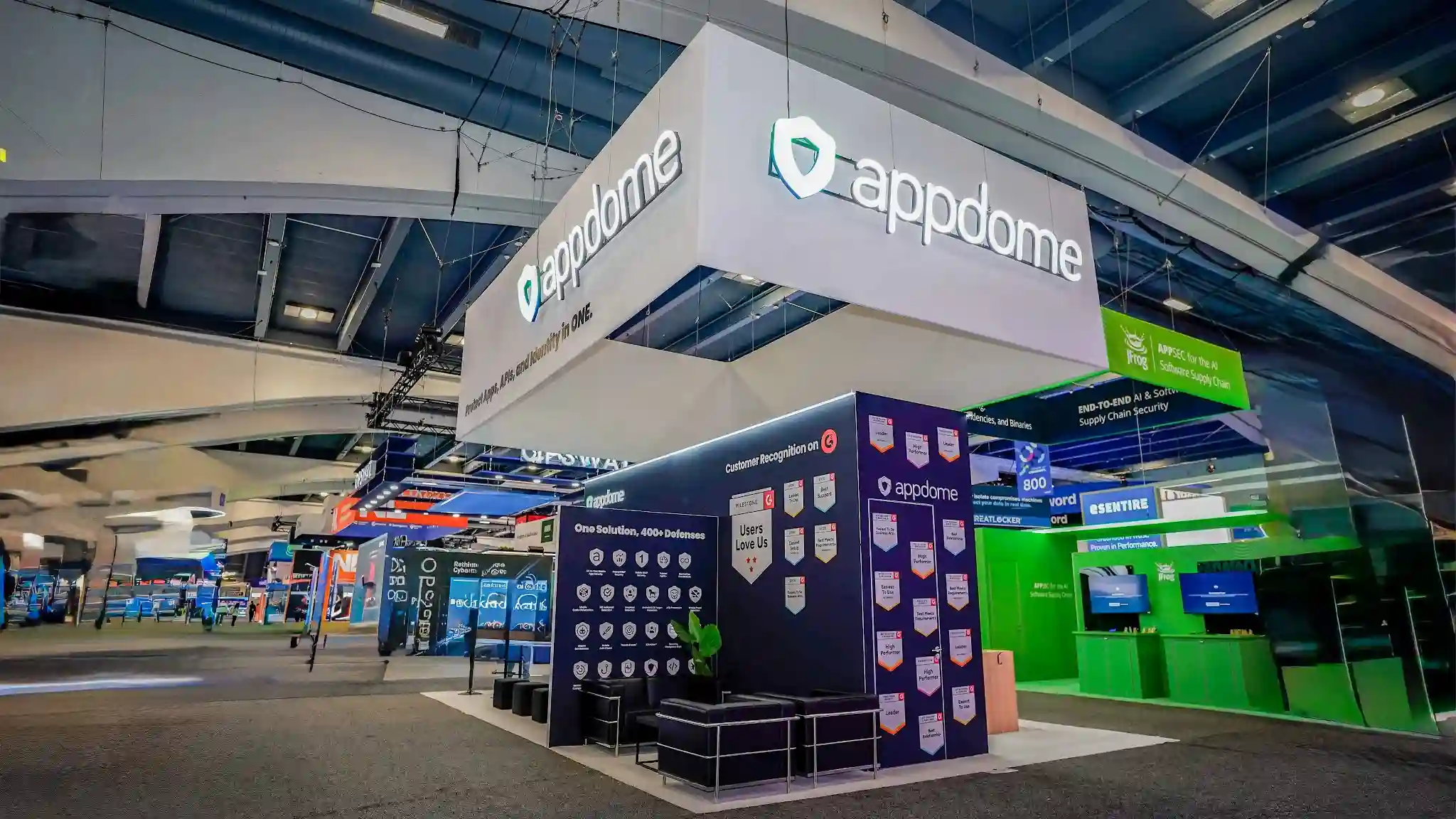 Low-angle three-quarter corner shot of the Appdome 20x30 ft island booth at RSA Conference 2026 taken from below and slightly to the left looking upward and inward showing the massive white internally-illuminated rectangular hanging sign dominating the upper half of the frame with appdome wordmark and teal green shield logo on the front face and Protect Apps APIs and Identity in ONE tagline on the side face suspended by steel cables from the convention ceiling, the dark navy backwall module approximately 20 feet wide by 10 feet tall displaying One Solution 400 Plus Defenses text with a grid of small white shield icons representing individual defense capabilities and a dedicated Customer Recognition on G2 awards panel with the G2 red logo and Users Love Us badge and approximately 15 to 20 G2 award badge icons arranged vertically including Best Support High Performer and Best Results categories, a tall freestanding column tower on the right with Appdome logo and additional G2 badges, black cushioned low lounge chairs visible in the lower foreground interior, a thin cyan teal LED accent strip along the top edge of the main display wall, white raised platform flooring, a small green potted plant adding organic warmth, JFrog bright neon green booth with APPSEC for the AI Software Supply Chain messaging visible to the right, eSentire blue and white booth signage visible in the background, dark convention hall ceiling with blue tinted ambient lighting