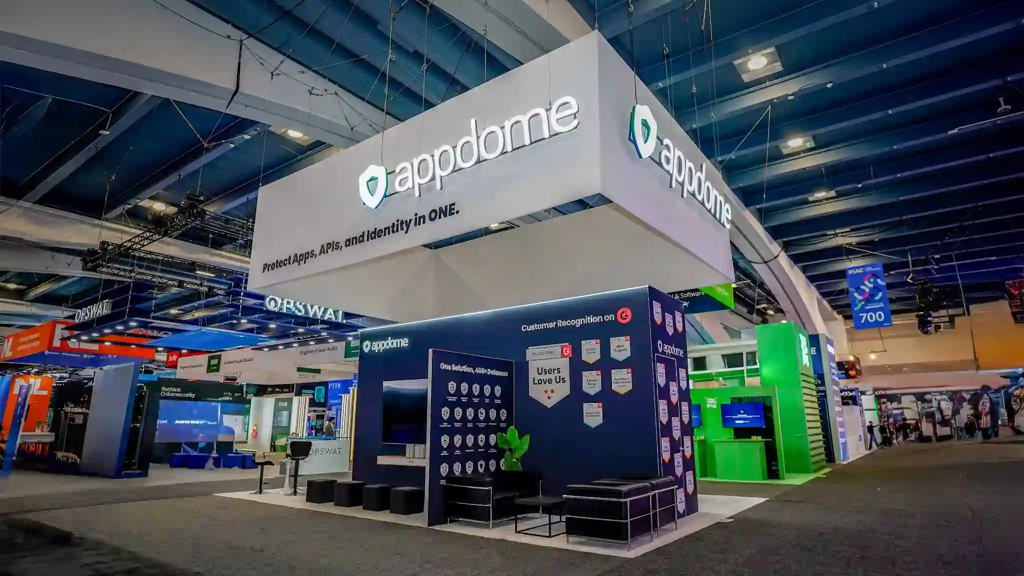 Wide establishing shot of the Appdome island exhibit presence at RSA Conference 2026 in the South Hall of Moscone Center San Francisco showing the booth environment on the convention show floor with overhead hanging signage prominently visible against the dark convention hall ceiling, white raised platform flooring creating a clean boundary against the dark charcoal convention carpet, structural columns and display panels with branding and campaign messaging, bar-height counter with stools for product demonstrations and visitor engagement, convention hall ambient lighting and neighboring exhibitor booths visible in the background including NETSCOUT booth with teal and green branding, aisle markers and directional signage indicating the South Hall layout, the overall premium enterprise-grade trade show exhibit aesthetic