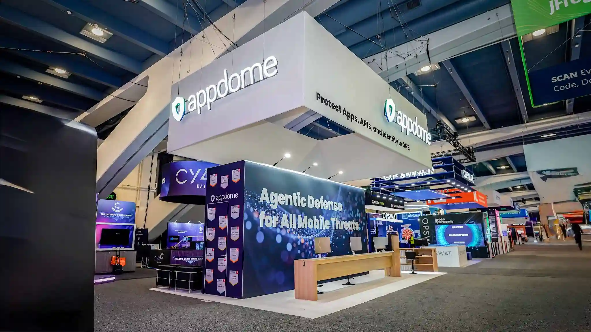 Blond wood demo counter with three monitor stations and gooseneck mics in front of the Agentic Defense for All Mobile Threats constellation wall at the Appdome booth, RSA 2026