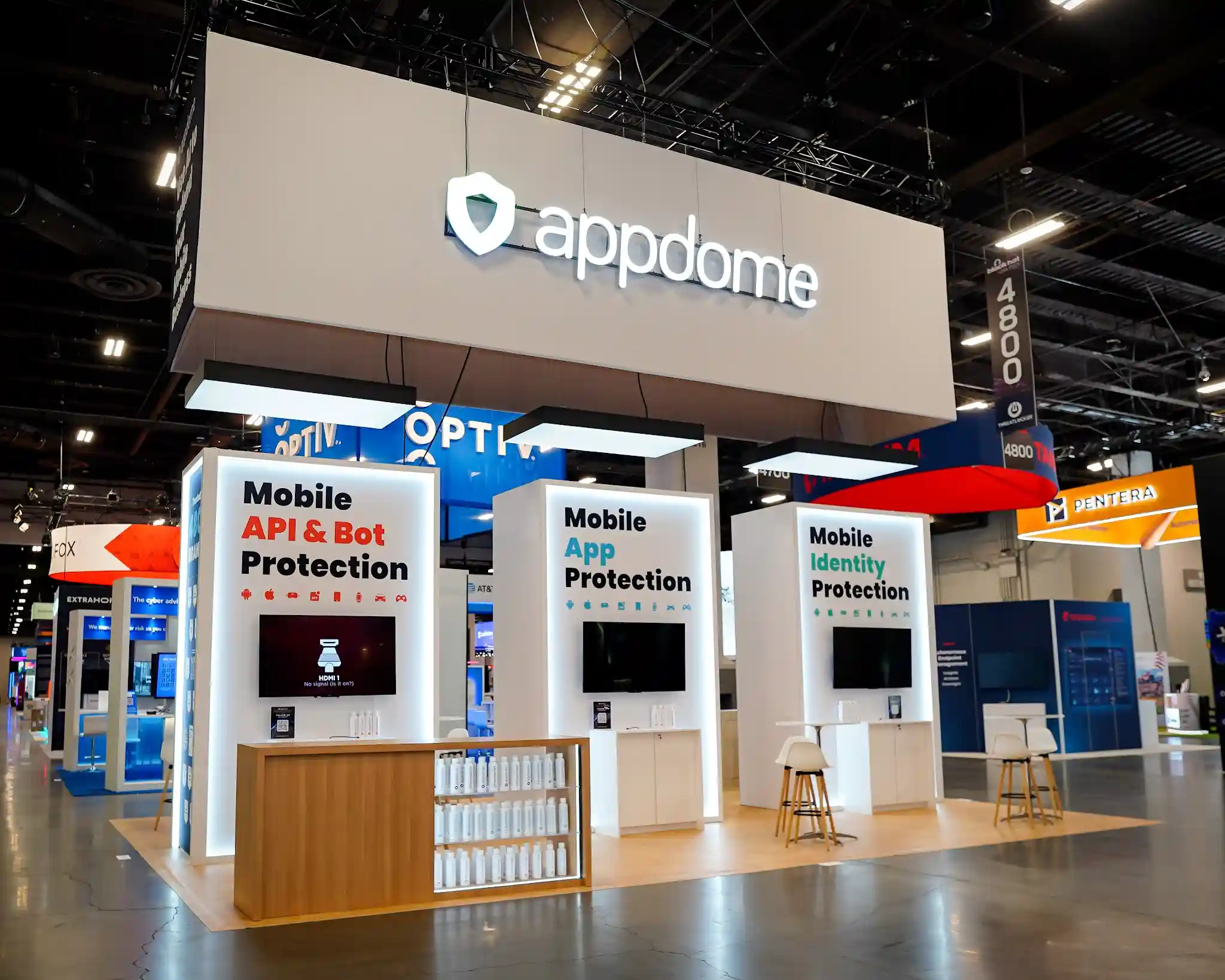 Three-quarter right-front view of Appdome island exhibit at Black Hat USA 2025 showing three white product towers with recessed monitors displaying interactive content, central natural wood bar counter with rows of white branded merchandise bottles on open lower shelving, white pedestal tables and Scandinavian barstools positioned at each tower, LED edge lighting along tower profiles, and neighboring Optiv booth visible behind the clean gallery-like exhibit layout