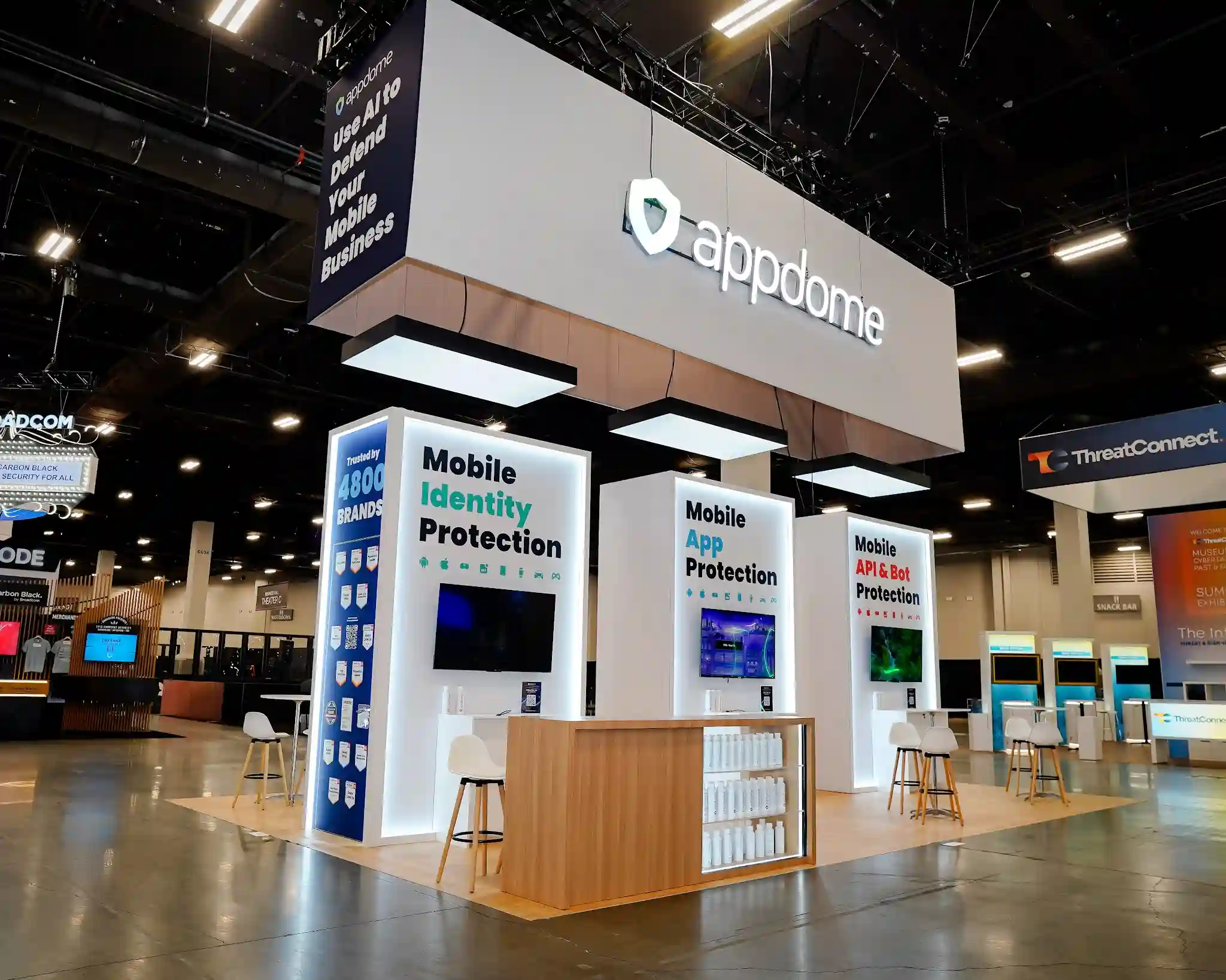 Three-quarter front view of Appdome 20x30 custom island exhibit at Black Hat USA 2025 showing massive white overhead canopy with illuminated appdome shield logo, dark navy Use AI to Defend Your Mobile Business hanging sign, three white product towers with LED edge lighting for Mobile Identity Protection Mobile App Protection and Mobile API and Bot Protection, central natural wood bar counter with white branded merchandise bottles, white Scandinavian barstools, raised natural wood platform flooring, and navy Trusted by 4800 BRANDS social proof column at Booth 4746 in the Mandalay Bay Convention Center Business Hall