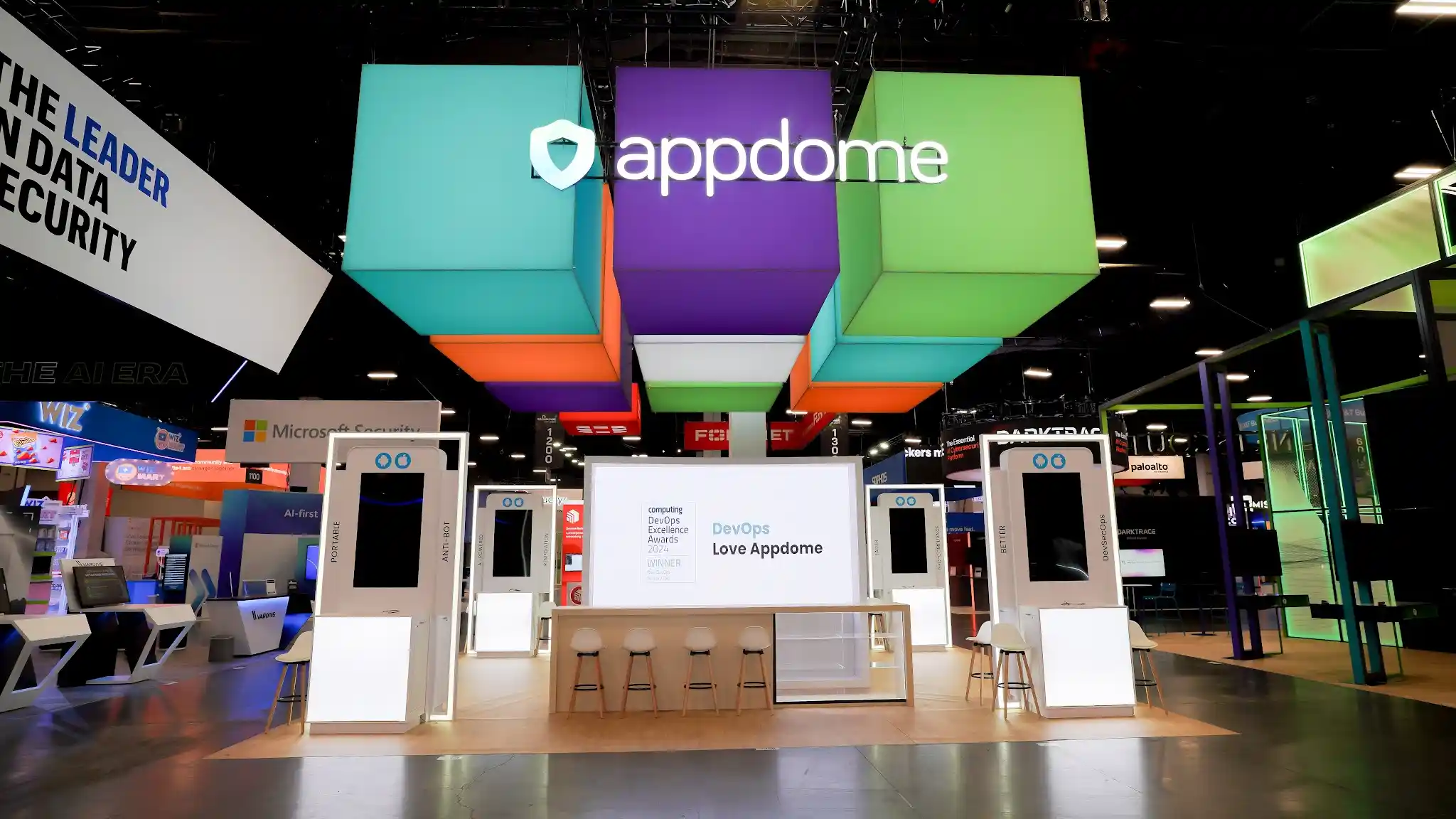 Straight-on frontal view from aisle of Appdome island exhibit showing DevOps Love Appdome messaging on central display panel, horizontal colorful cube arrangement in purple turquoise lime green coral orange and white spelling out the brand name, symmetrical white MOBILE LINK kiosk stations flanking the entrance, natural wood flooring, and structured engagement zones at Booth 1200 Black Hat USA 2024