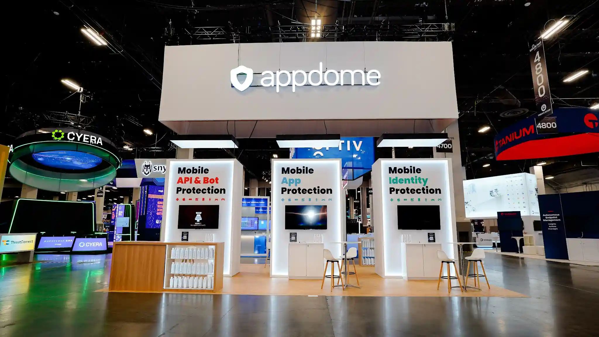 Wide symmetrical frontal view of Appdome 20x30 island exhibit at Black Hat USA 2025 from distance showing full booth in context with neighboring cybersecurity booths including Cyera circular booth Tanium and Optiv, three white product towers evenly spaced with LED edge lighting, natural wood bar counter visible at center, white overhead canopy with illuminated appdome logo glowing above, and clean gallery-like aesthetic standing out against the crowded Business Hall show floor at Mandalay Bay Convention Center
