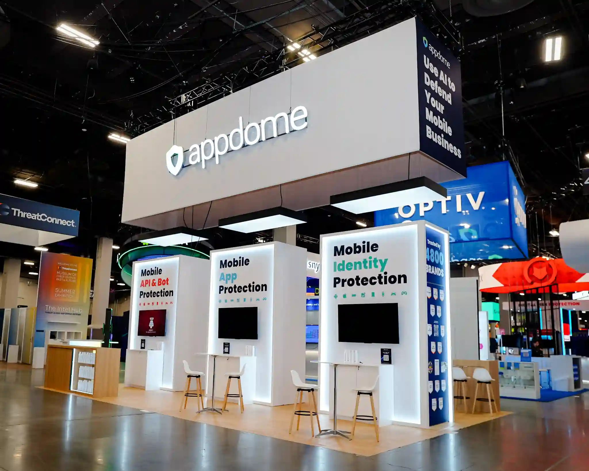 Front-facing view of Appdome 20x30 island exhibit at Black Hat USA 2025 showing three white product towers with LED edge lighting from opposite angle, white overhead canopy with illuminated appdome shield logo, neighboring ThreatConnect and Optiv booths visible for context, natural wood flooring platform creating a defined island footprint, and Scandinavian barstools positioned at each product station for one-on-one technical consultations in the Mandalay Bay Convention Center Business Hall