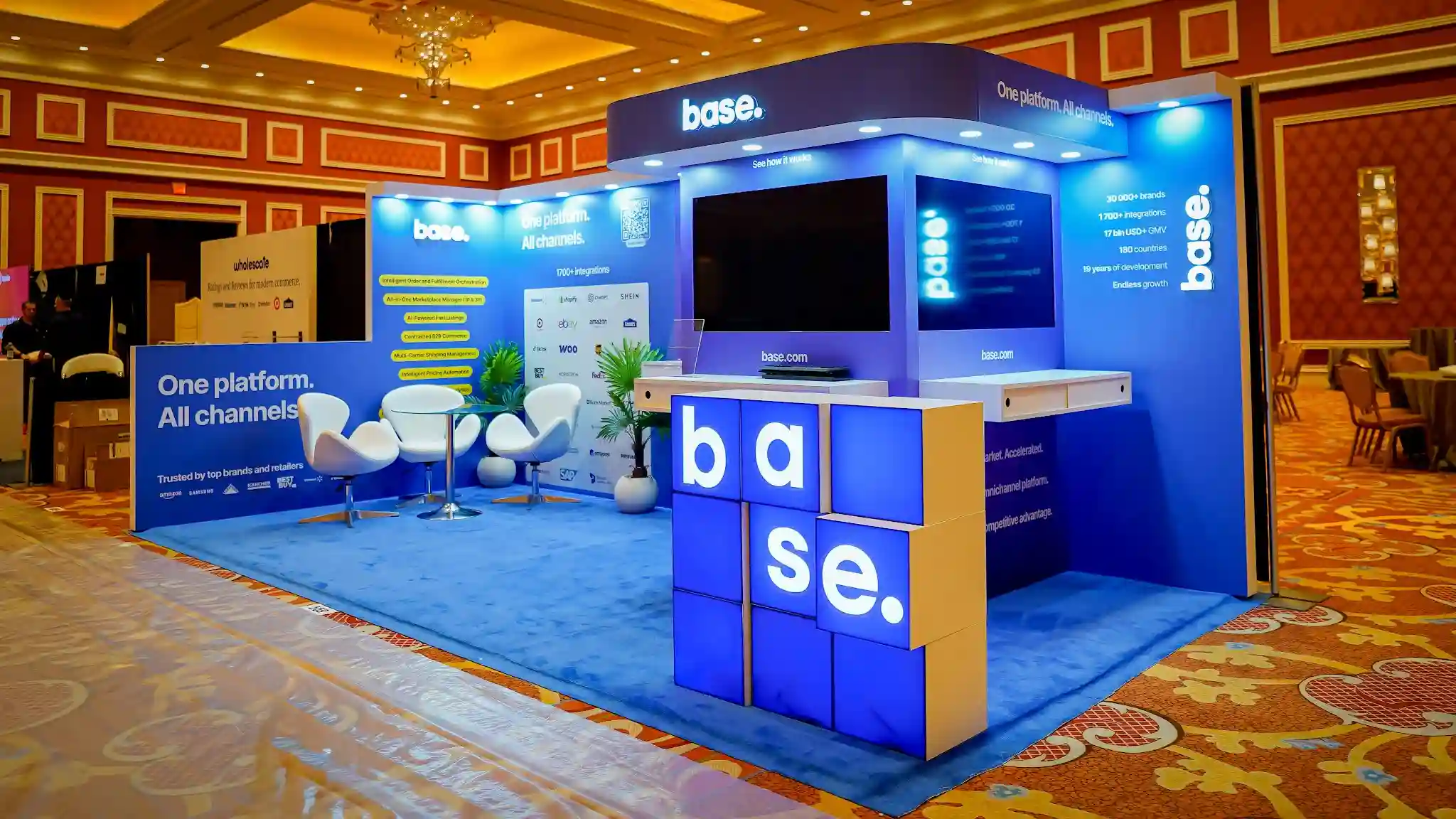 Front-center full view of the Base booth at Prosper Show 2026 showing the complete layout with left back wall featuring One platform All channels branding and Trusted by top brands and retailers section with Amazon Samsung Adidas Walmart logos, center demo area with flat-screen monitors, raised kiosk tower with curved canopy and base dot header, freestanding illuminated base letter-cube installation at front right corner with blue and beige cubes, three white egg pod chairs with glass table in lounge, tropical plant, yellow feature badge pills for AI-Powered Fast Listings and Intelligent Order Fulfillment, blue carpet flooring, LED downlights, warm Wynn ballroom lighting overhead