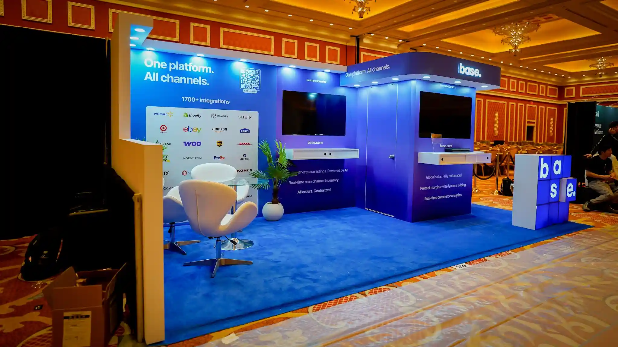 Front-center slightly right angle of the Base 10x20 ft booth at Prosper Show 2026 showing the full booth from a closer perspective with the left wall One platform All channels branding and 1700 plus integrations partner logo grid clearly visible, center flat-screen monitor between left wall and right tower, right kiosk tower with curved canopy header and base dot branding, base letter-cube installation prominent at the right corner with blue and beige cube faces, white egg pod lounge chairs and glass table in the center, tropical plant adding green accent, blue carpet flooring, recessed LED downlights, warm Wynn ballroom chandelier lighting overhead creating golden warmth contrast