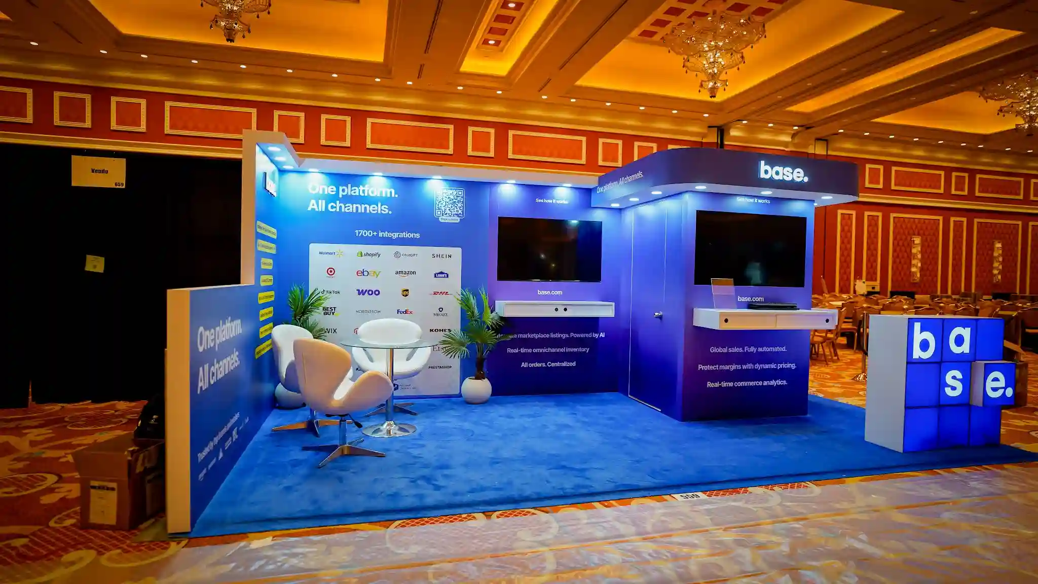 Front-left wide-angle perspective of the Base 10x20 ft inline booth at Prosper Show 2026 showing the full booth layout from the left flat-wall section with One platform All channels headline and 1700 plus integrations partner logo grid to the right elevated kiosk tower with curved canopy header and base dot branding, dual flat-screen monitors, white egg pod lounge chairs with glass round table and tropical plant in the center lounge zone, freestanding illuminated base letter-cube installation at the far right corner, recessed LED downlights along top edges of both wall sections, blue carpet flooring, warm Wynn ballroom ambient lighting from chandeliers overhead