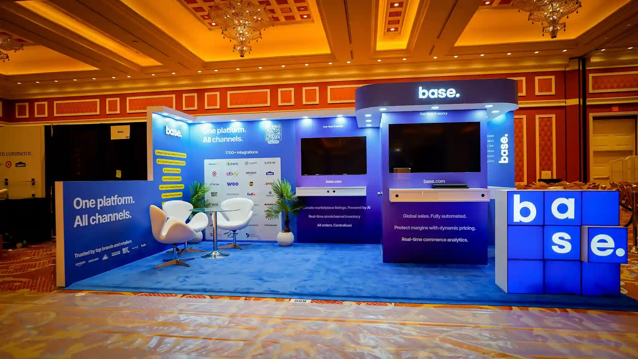 Wide front-facing establishing shot of the complete Base 10x20 ft inline booth at Prosper Show 2026 Booth 559 at The Wynn Las Vegas showing the full booth footprint with vivid royal cobalt blue wall panels and blue carpet flooring, raised right-side kiosk tower with curved overhead canopy header featuring backlit base dot logo in white, dual flat-screen monitors mounted on center and right wall sections with white shelf counters beneath for product demonstrations, freestanding illuminated three-dimensional base letter-cube installation at the front right corner with alternating blue and beige cube faces spelling b a s e in large white letters, left back wall panel with One platform All channels headline in large white text and 1700 plus integrations partner logo grid showing Amazon Walmart Shopify eBay TikTok ChatGPT SHEIN FedEx DHL Best Buy Nordstrom Kohls and more, yellow pill-shaped feature badges for AI-Powered Fast Listings and Intelligent Order Fulfillment Orchestration, white egg pod lounge chairs with glass-top round table and tropical potted plant in the lounge area, recessed LED downlight spotlights along top edges of wall panels creating bright ambient illumination, warm golden ambient light from The Wynn ballroom ornate coffered ceiling with chandeliers contrasting with cool blue booth lighting