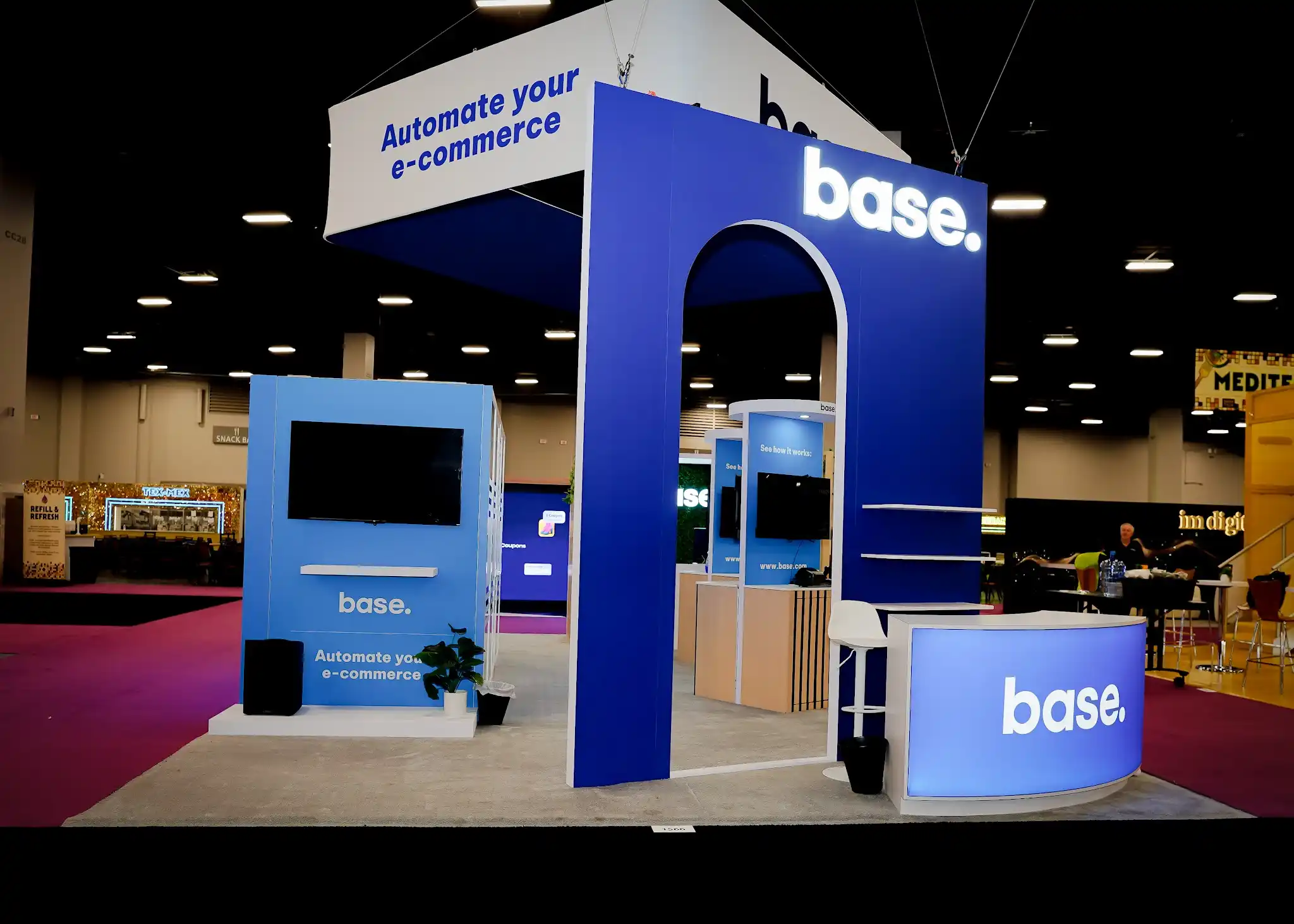 Angled perspective of the Base booth at Shoptalk 2025 showing three sides of the island exhibit with the tall dark navy arch structure in the center rear featuring a rounded white archway portal and base logo in white illuminated letters at top, curved gradient blue to purple backlit reception desk in the right foreground with base branding, sky blue kiosk unit on left with large flat screen monitor and base Automate your e-commerce text, overhead white hanging sign with dark blue text visible above, white raised platform flooring, green potted plants at corners, white high-top barstool behind desk, dark convention ceiling with recessed grid lighting