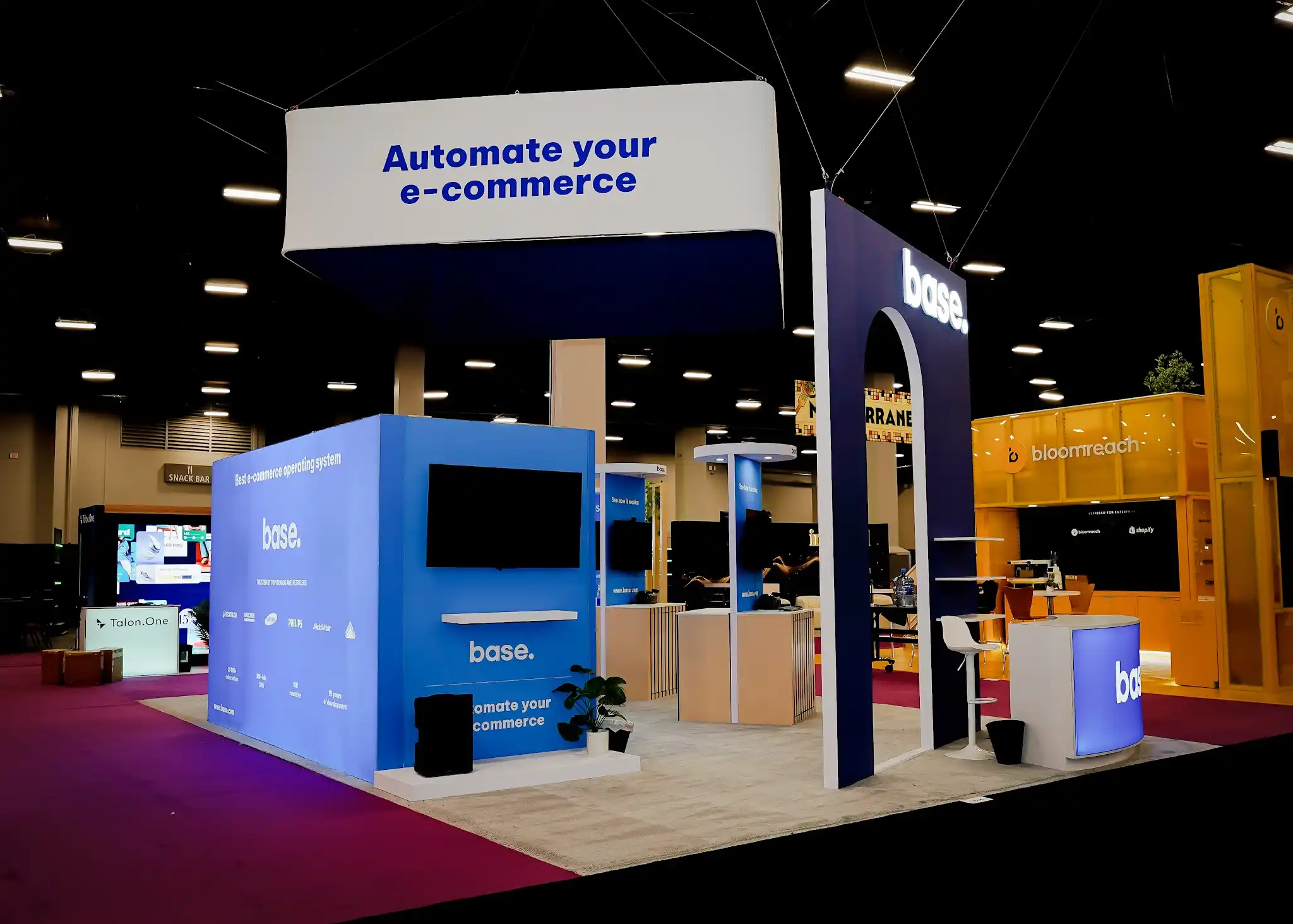 Wide front-left view of the Base 20x30 ft island booth at Shoptalk 2025 showing the large white integration showcase wall panel displaying base logo in dark blue followed by Best e-commerce operating system tagline and Trusted By Top Brands and Retailers text with client logos including Decathlon Karcher Samsung Philips and MediaMarkt plus statistics reading 30000 plus online sellers and 16 plus bln GMV and 180 countries and 19 years of development, tall dark navy arch structure with white arch portal on the right side, Bloomreach bright yellow booth visible adjacent, Aisle 1500 hanging banner in background, Content Square red booth in far background, light gray raised platform flooring, deep magenta purple convention carpet in aisles, small potted green plants at base of panels