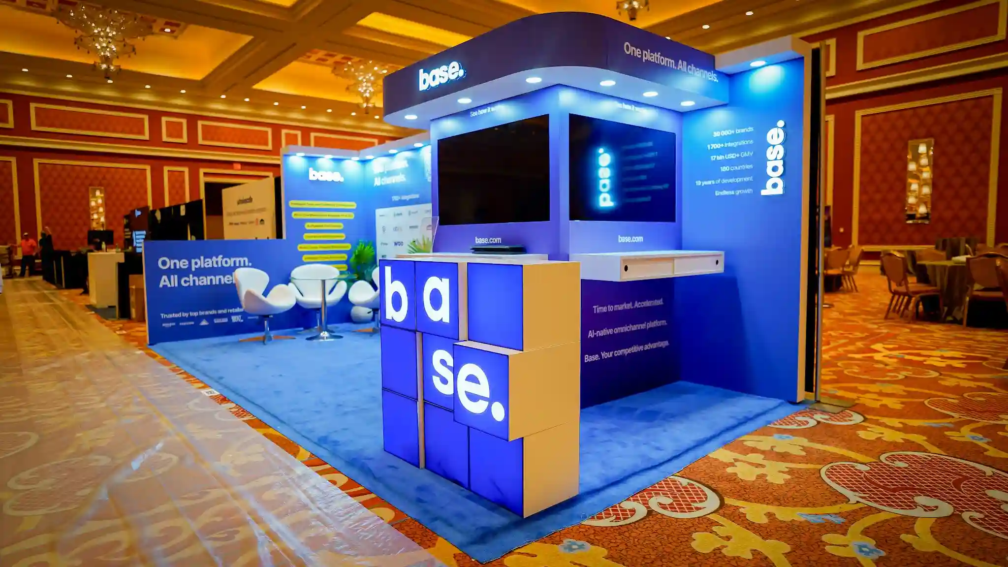 Corner diagonal angle of the Base booth at Prosper Show 2026 showing the full island configuration with the illuminated base letter-cube installation prominent in the foreground, right kiosk tower with curved canopy behind it, left wall visible in the distance, stats column on right panel showing 30000 plus brands 1700 plus integrations 17bn USD plus GMV 180 countries, blue carpet, warm ballroom lighting