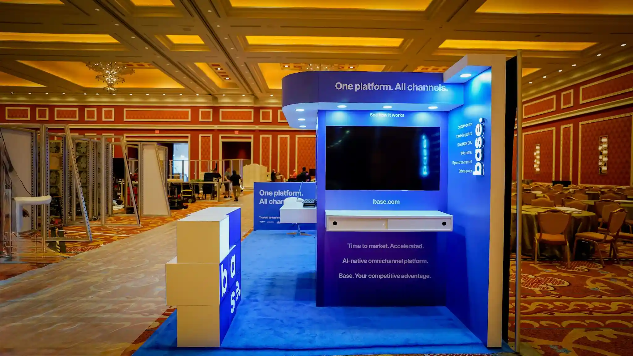 Close-up front-right view of the Base booth kiosk tower section at Prosper Show 2026 during setup showing the raised tower structure with dramatic curved rounded overhead canopy header displaying base dot logo in backlit white against dark navy panel, One platform All channels sub-header text, See how it works call-to-action, large flat-screen monitor mounted centrally with white shelf counter beneath for demonstrations, right-side vertical panel showing stats column with 30000 plus brands 1700 plus integrations 17bn USD plus GMV 180 countries 19 years of development Endless growth, base dot letter-cube installation partially assembled in left foreground, blue carpet flooring, construction workers and ladders visible in background indicating pre-show setup, warm Wynn ballroom wooden ceiling overhead
