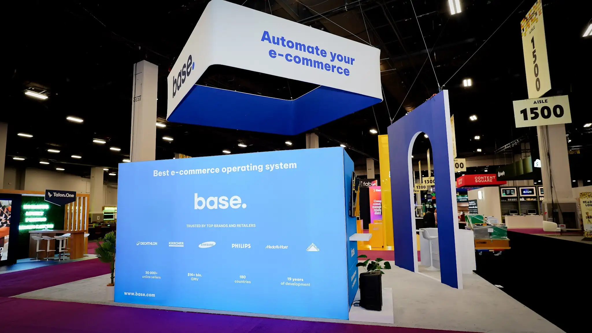 Upward angle focusing on the Base overhead hanging sign with Automate your e-commerce in dark blue italic text on white background, blue top soffit panel, steel suspension cables, navy arch in profile, show floor booths visible in distance