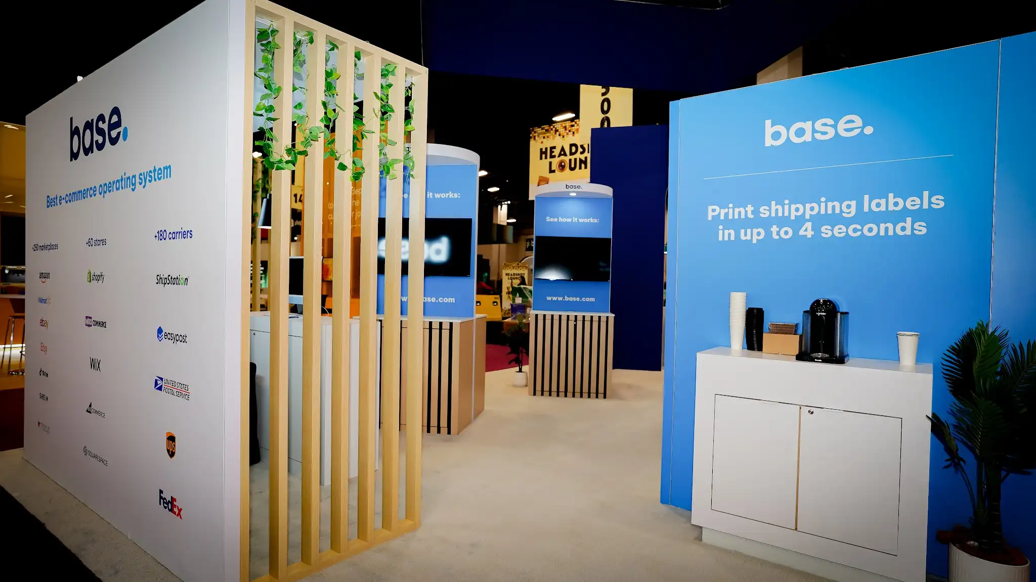 Interior view of the Base booth at Shoptalk 2025 looking toward the back showing the decorative vertical wood slat partition with cascading green ivy plants on the left, white integration wall panel on the far left displaying base logo Best e-commerce operating system text and partner integration grid with Amazon Walmart eBay Shopify WooCommerce USPS UPS FedEx logos in three columns, bright blue side panel reading Print shipping labels in up to 4 seconds with white storage cabinet and coffee machine refreshment station below, cylindrical demo kiosk towers with blue panels and wood-slat lower wraps in center background, light gray exhibition flooring throughout, dark convention ceiling, neighboring booths visible in far background