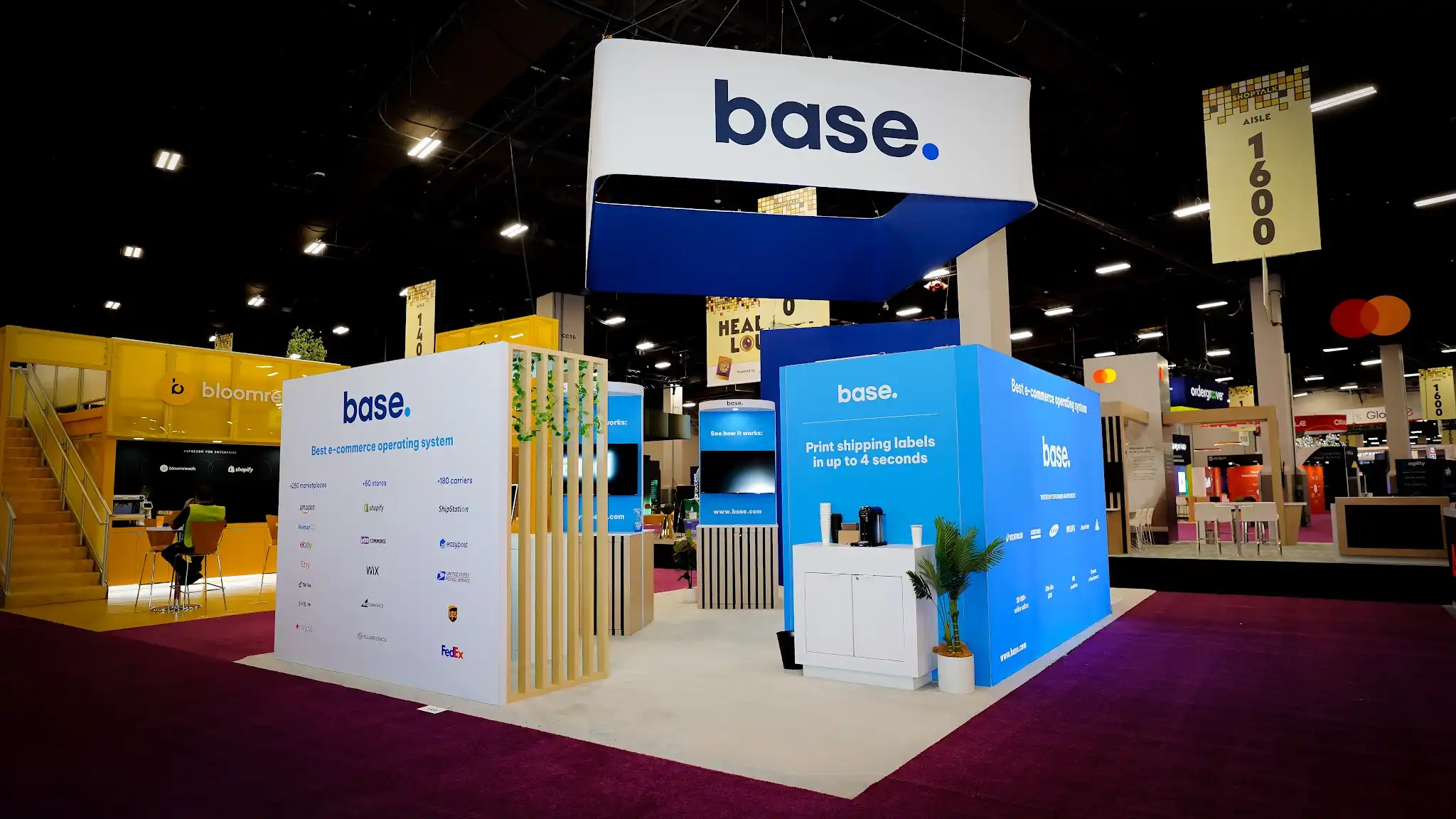 Wide establishing shot of the complete Base 20x30 ft island exhibit at Shoptalk 2025 in the Mandalay Bay Convention Center showing the full front facade with the large rectangular overhead hanging sign displaying Automate your e-commerce in dark blue italic text on white background, decorative vertical wood slat partition with cascading green ivy plants in the center, tall dark navy arch structure with rounded white portal and base logo in white letters at top, integration showcase wall on the left displaying marketplace store and carrier partner logos, raised white platform flooring contrasting with the deep magenta purple convention carpet, white bar stools, cylindrical demo kiosks visible in interior, green plants throughout, neighboring exhibitor booths in background, dark black convention ceiling with rows of recessed LED strip lighting