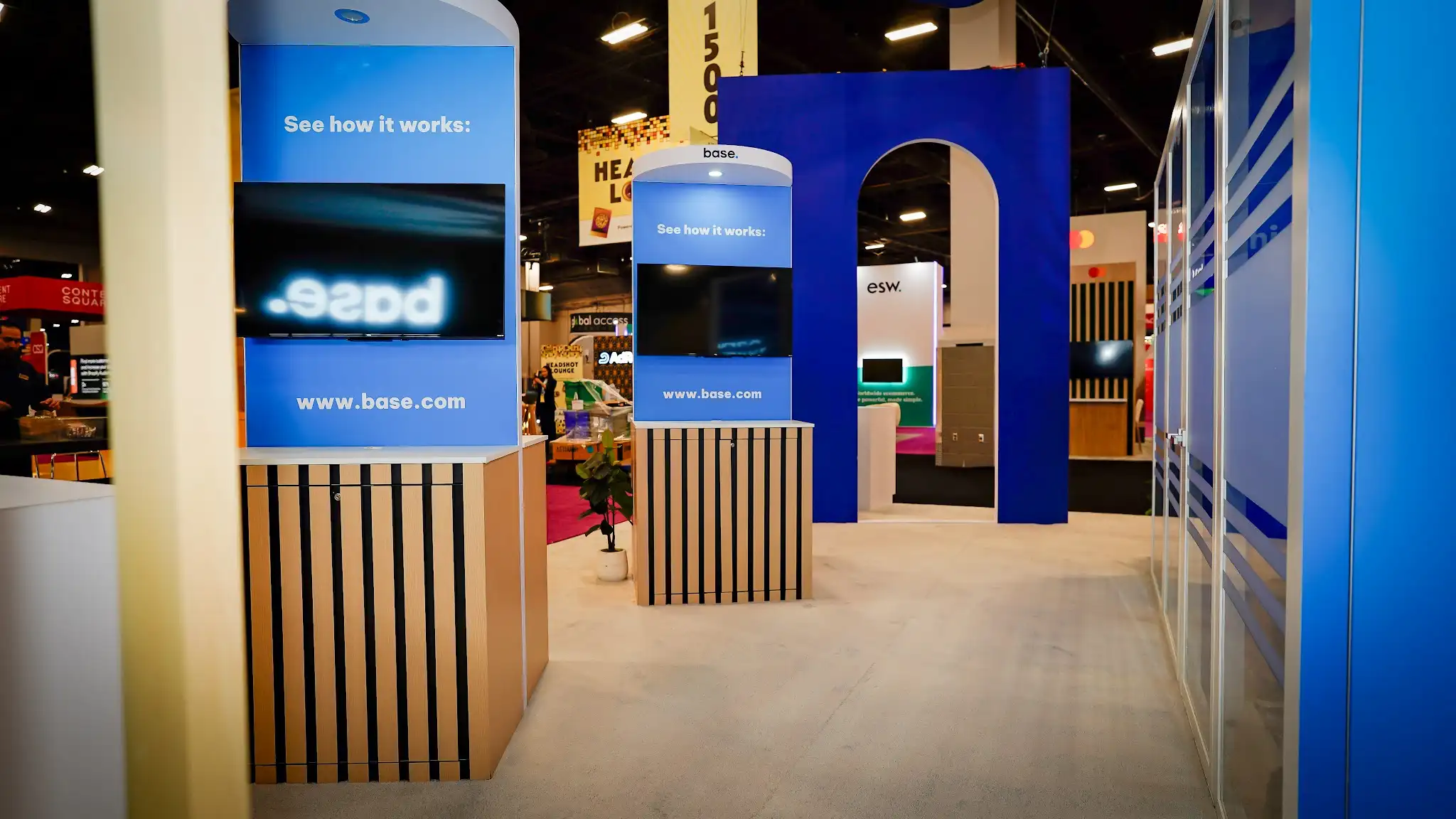 Interior corridor view of the Base booth showing cylindrical demo kiosk towers with wood-slat lower wraps and blue See how it works panels with embedded monitors, navy arch structure in background, meeting rooms edge visible on right, white planters with plants