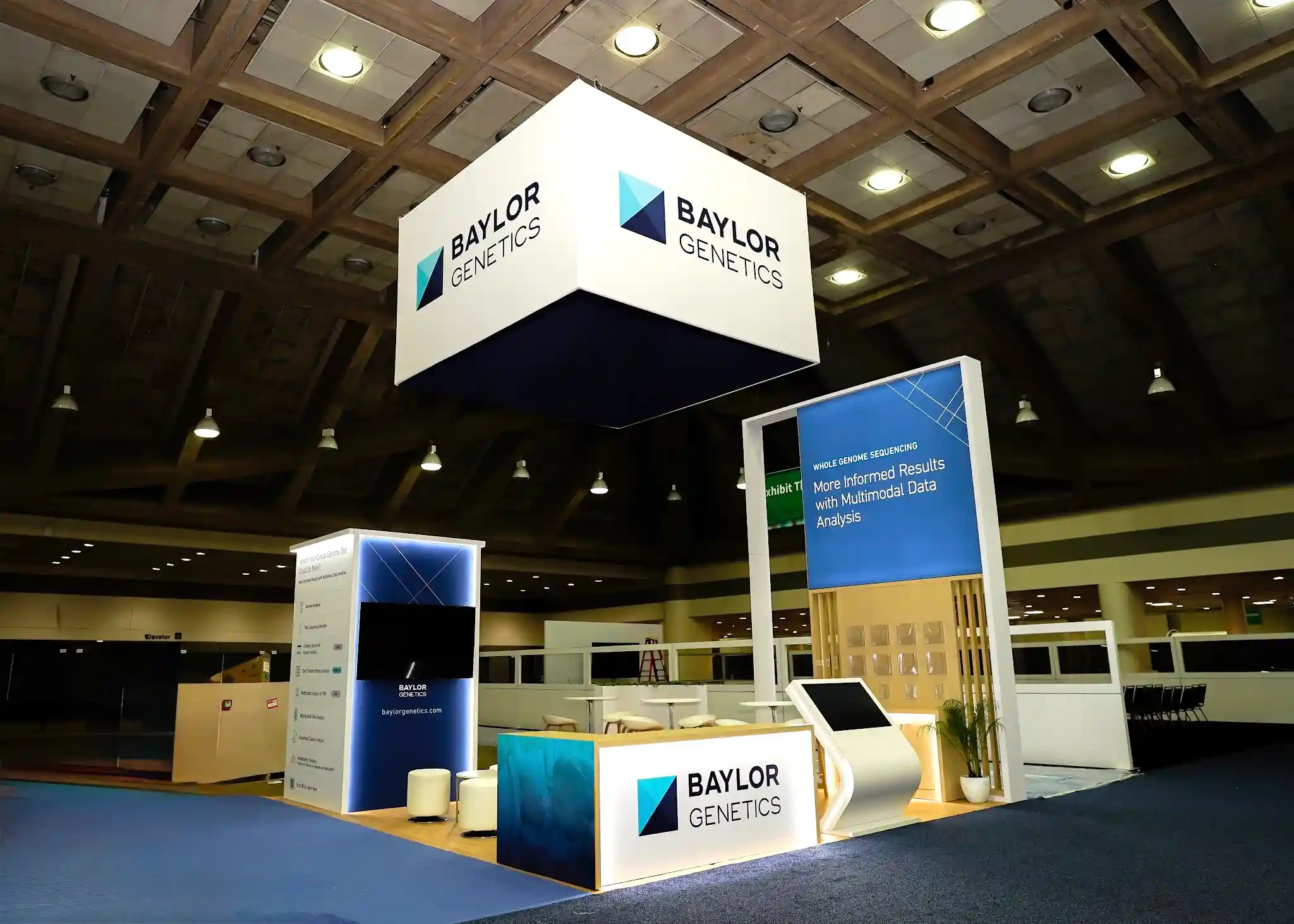 Baylor Genetics 20x20 custom island booth at ACMG 2026 featuring angled front view with overhead hanging sign, cylindrical tower display, white reception counter, WGS tower with natural wood slat cladding, turquoise accent panel, and curved ottoman seating at Baltimore Convention Center