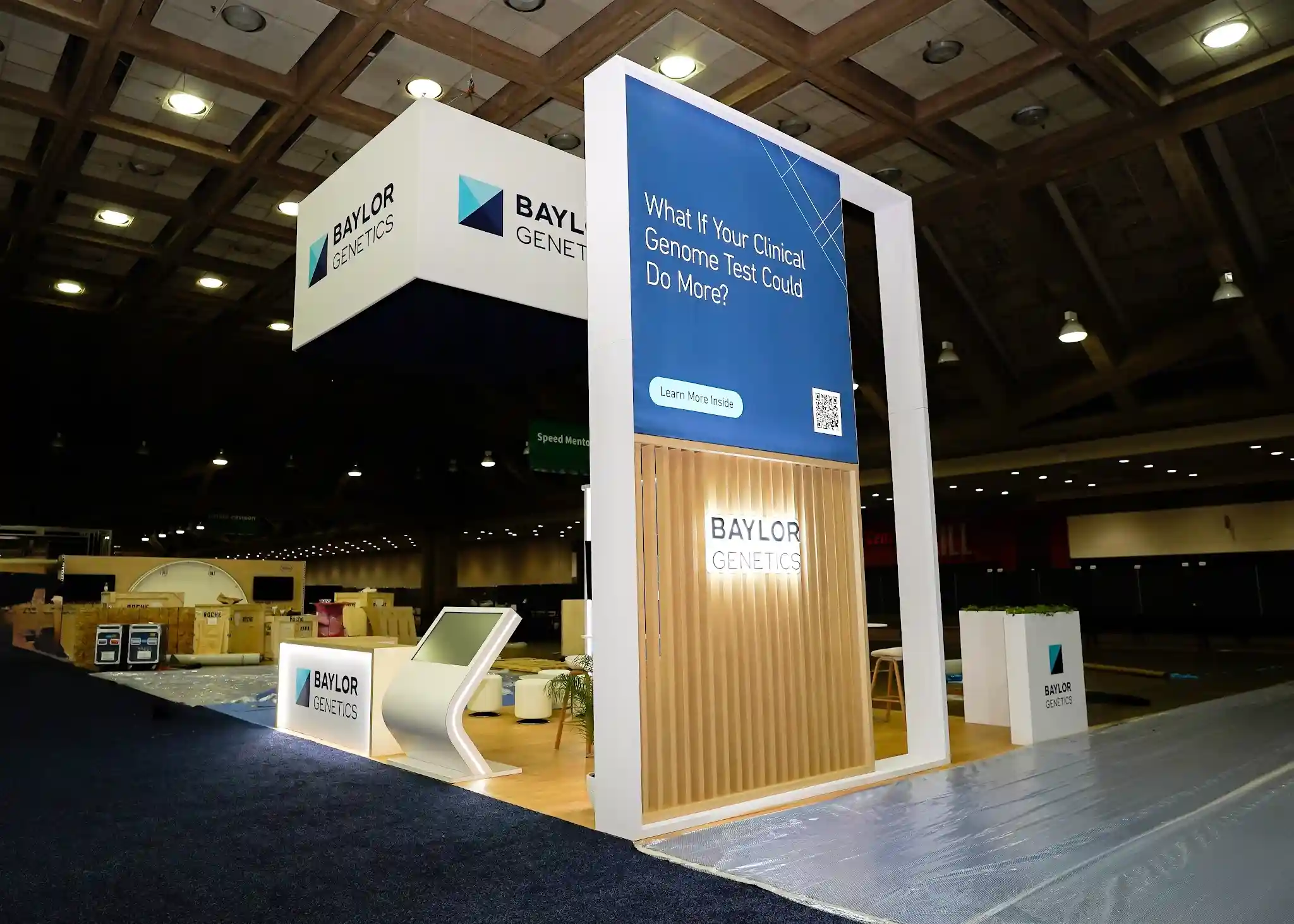 Corner angle of Baylor Genetics booth showing What If Your Clinical Genome Test interactive tower with large QR code, natural wood slat paneling, curved white reception desk with integrated tablet kiosk, and Baylor Genetics branding overhead